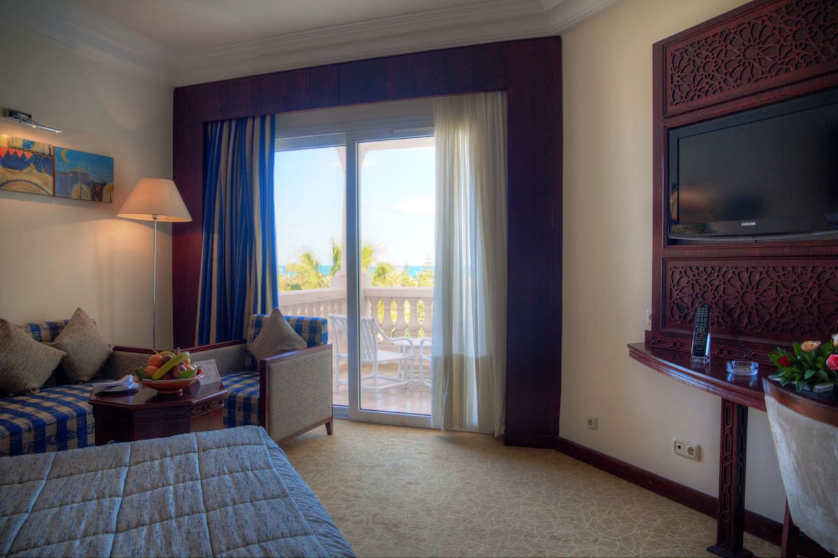 Une chambre d’hôtel avec un lit, un canapé, une table avec des fruits, une télévision et un balcon avec vue.