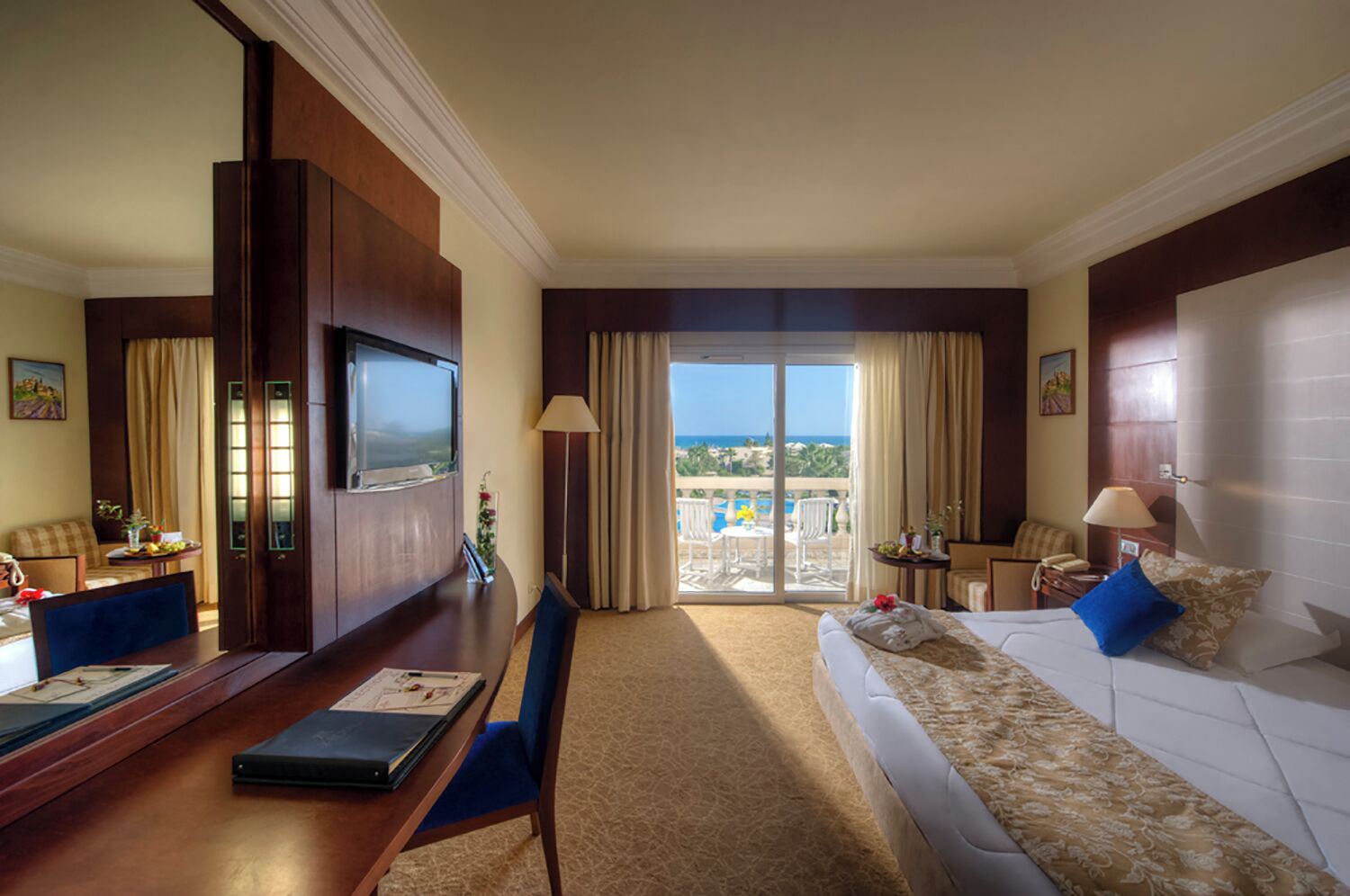 A hotel room with a large bed, a desk with a laptop, a TV, and a balcony with a view of the ocean.