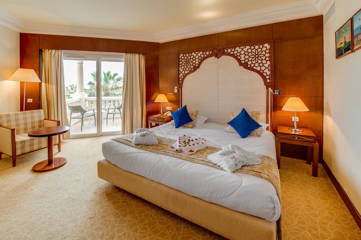 Une chambre d’hôtel avec un grand lit, deux tables de chevet avec des lampes, une petite table ronde et un balcon avec une chaise.