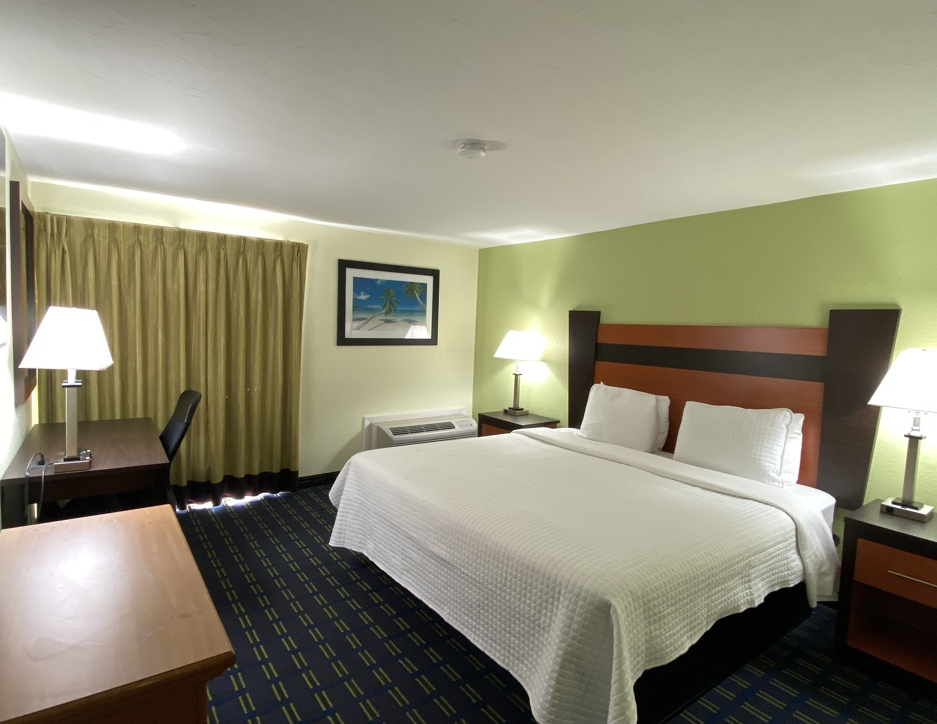 Habitación de hotel con cama, dos mesitas de noche, escritorio, silla, ventana con cortinas y aire acondicionado de pared.