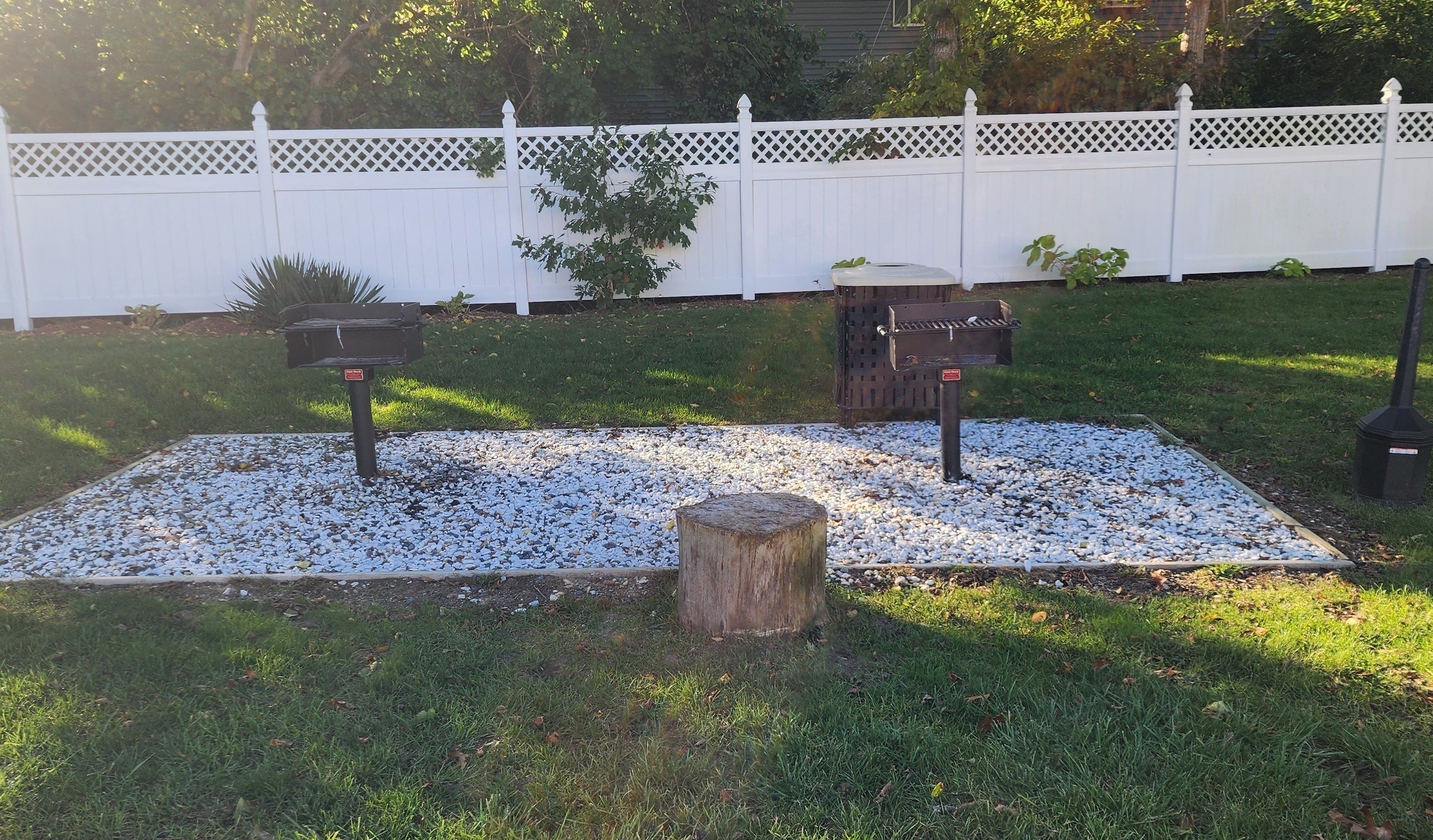 Un jardin avec une clôture blanche, deux barbecues et un tronc d’arbre.