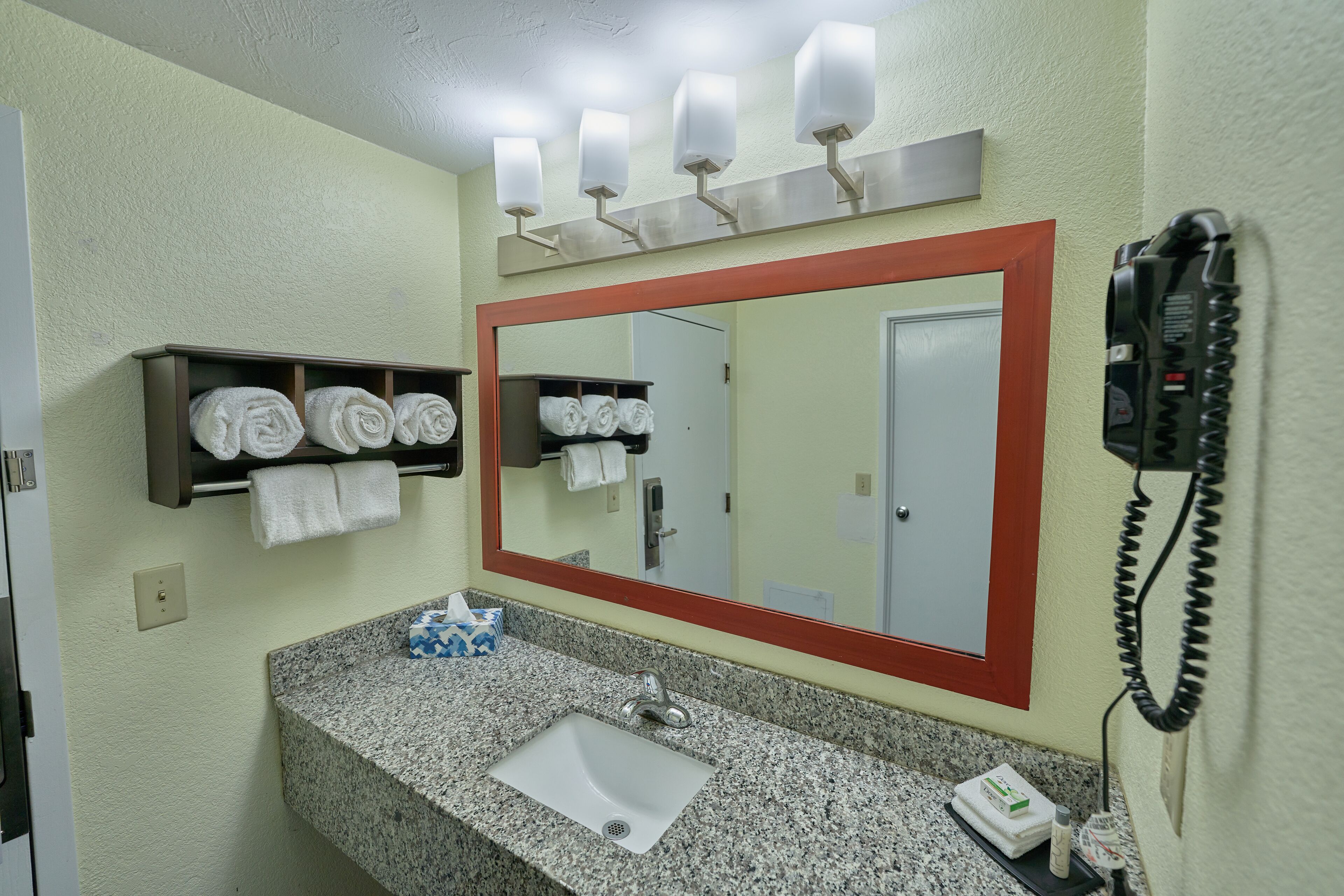A bathroom with a granite countertop sink, a mirror with a red frame, a towel rack with towels, and a telephone.
