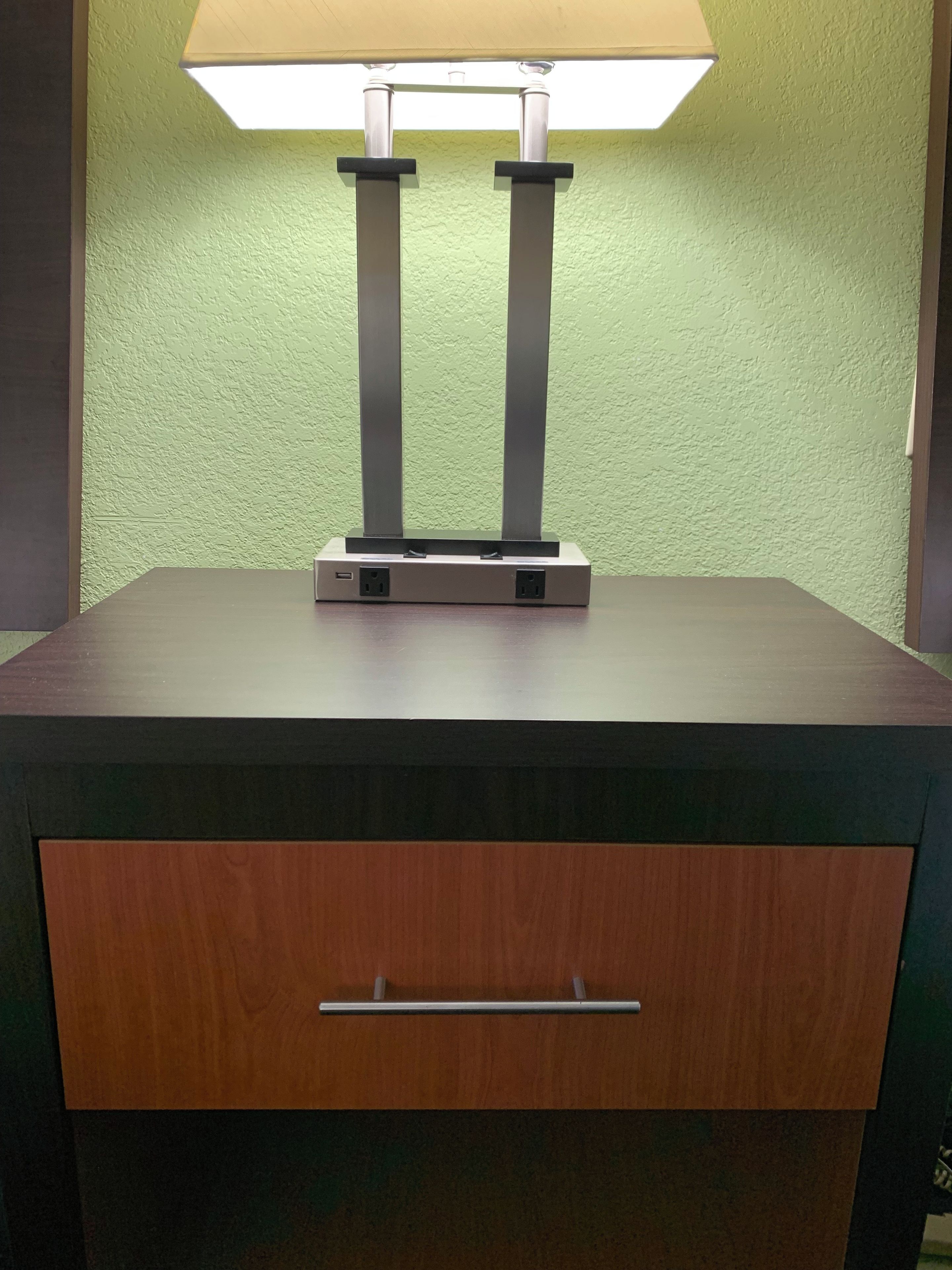 A table with a lamp and a power strip, a drawer, and a green textured wall.