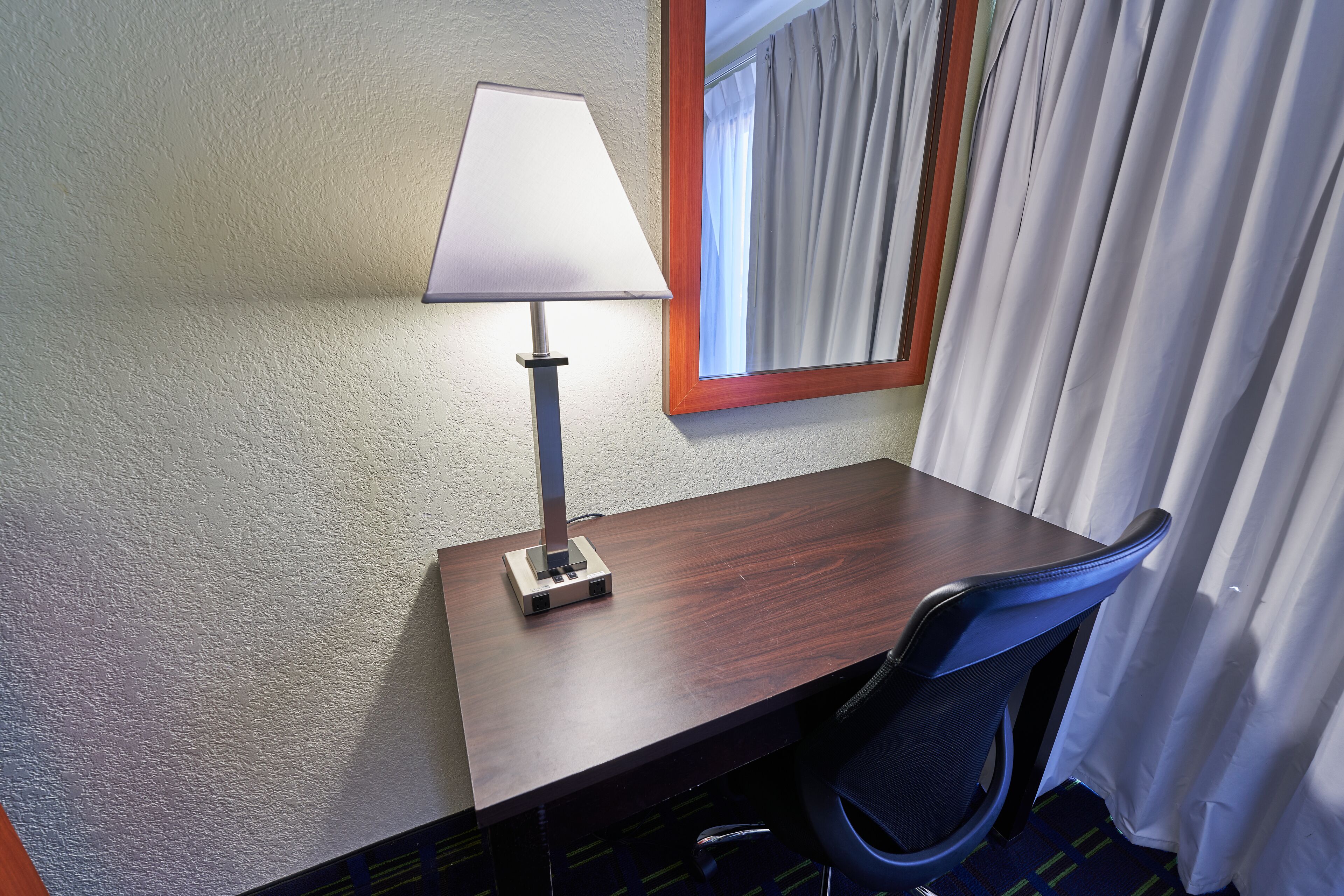 Une chambre d’hôtel avec un bureau en bois, une lampe, une chaise et une fenêtre avec des rideaux.