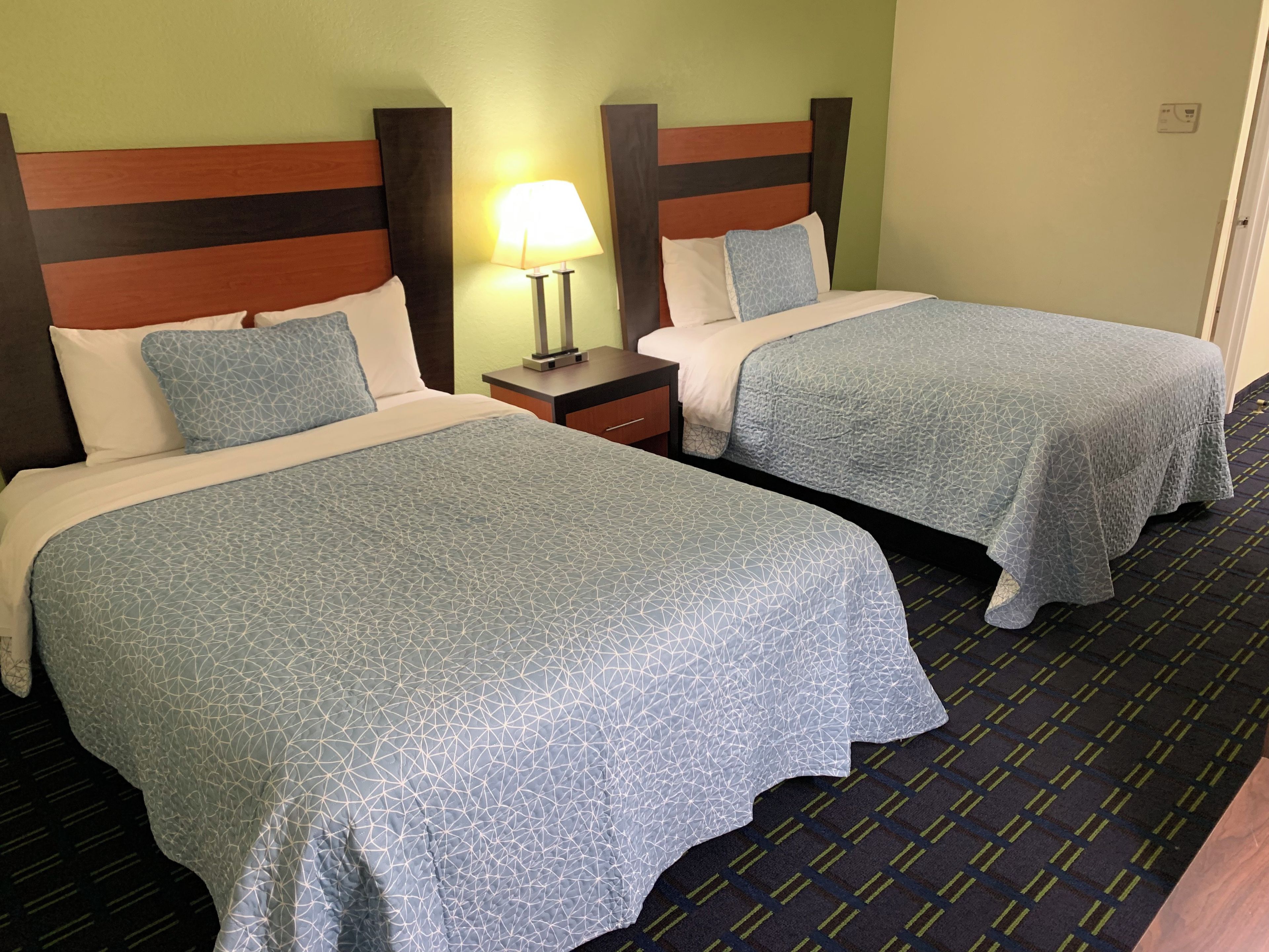 A hotel room with two beds, each with a patterned bedspread, a nightstand with a lamp, and a carpeted floor.