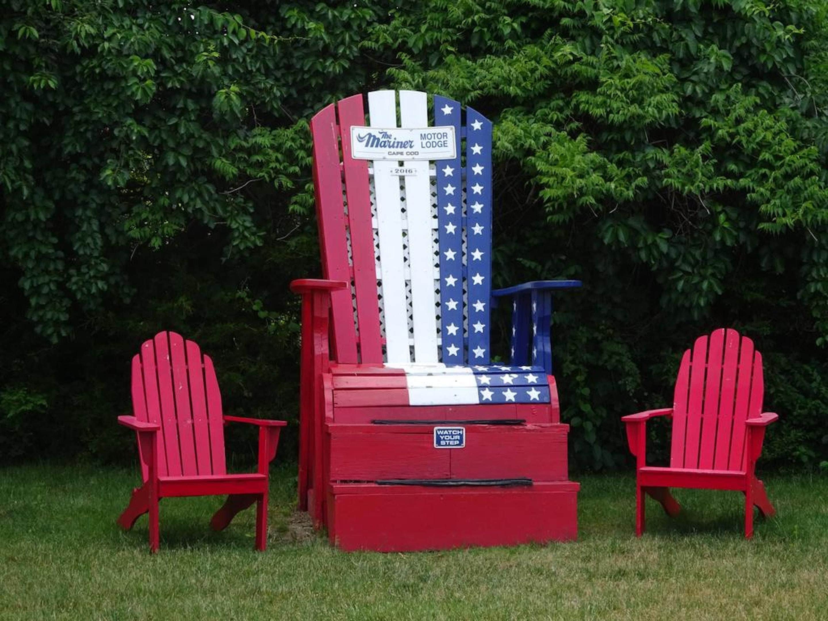 A red Adirondack chair with a flag design, a sign reading 'The Mariner Lodge Cape Cod,' and another chair beside it.