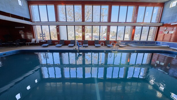 Une piscine intérieure à l’eau claire, entourée de chaises longues et offrant une vue sur l’extérieur grâce à de grandes fenêtres.