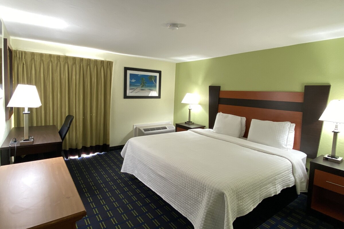 Ein Hotelzimmer mit einem Bett, zwei Nachttischen mit Lampen, einem Schreibtisch, einem Sessel, einem Fenster mit Vorhängen und einem Bild an der Wand.