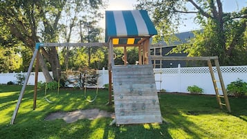 Eine Holzspielkonstruktion mit Rutsche und Kletterwand, umgeben von einem weißen Zaun und Grünpflanzen.