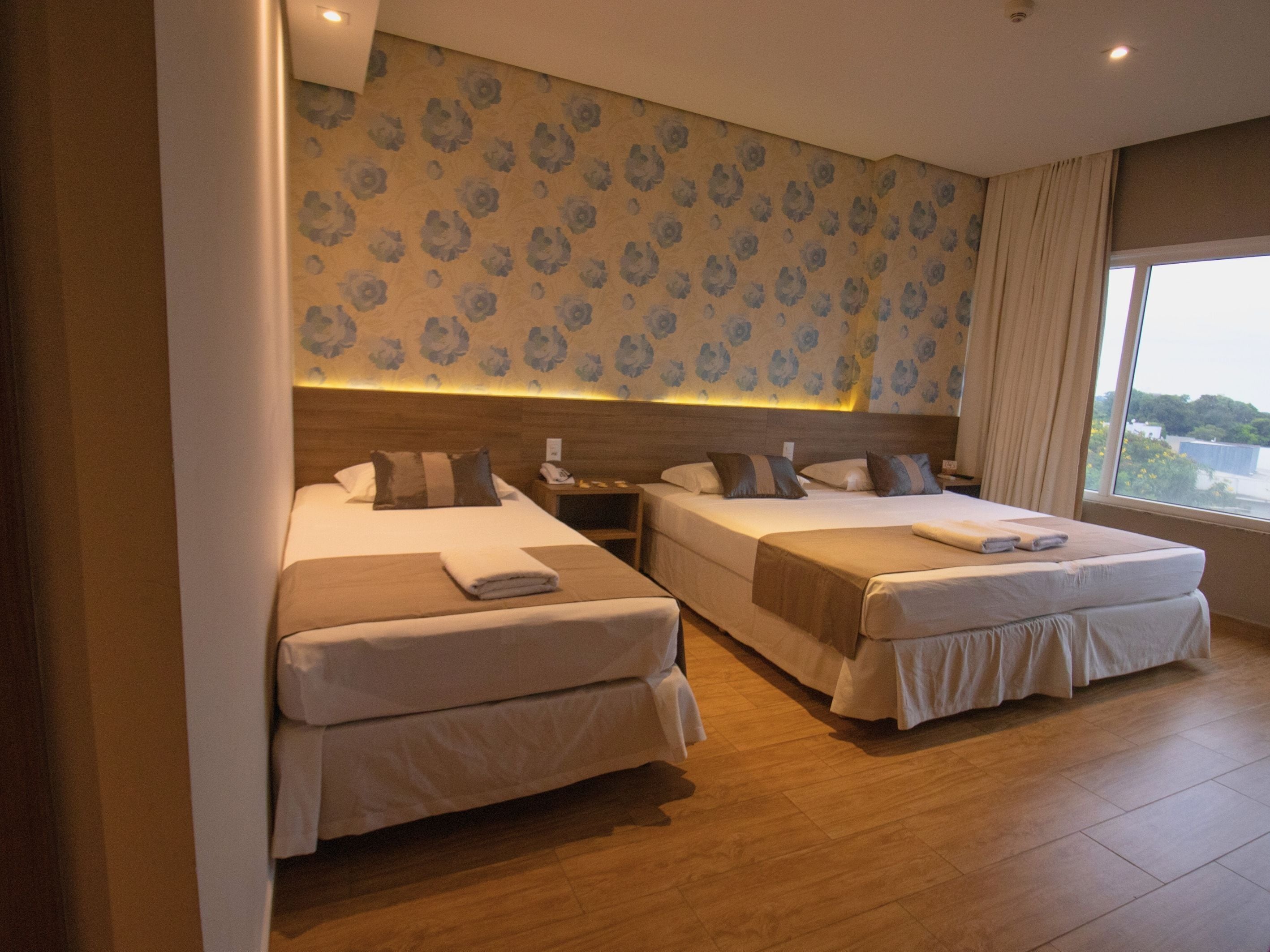 A hotel room with two beds, a nightstand, a lamp, and a large window with a view.