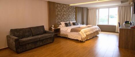 A modern hotel room with a large bed, a sofa, a desk, and a window with curtains.
