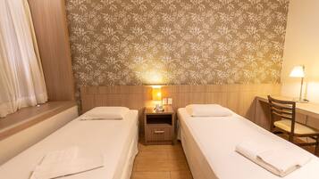 A hotel room with two single beds, a desk with a chair, a nightstand with a lamp, and a floral-patterned headboard.