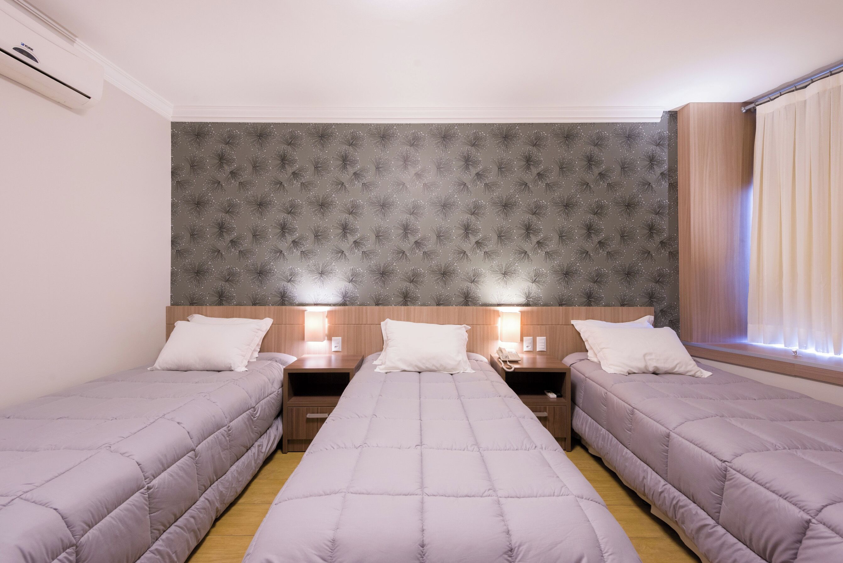 A hotel room with two single beds, a wooden headboard, a small desk with a lamp, and a window with curtains.