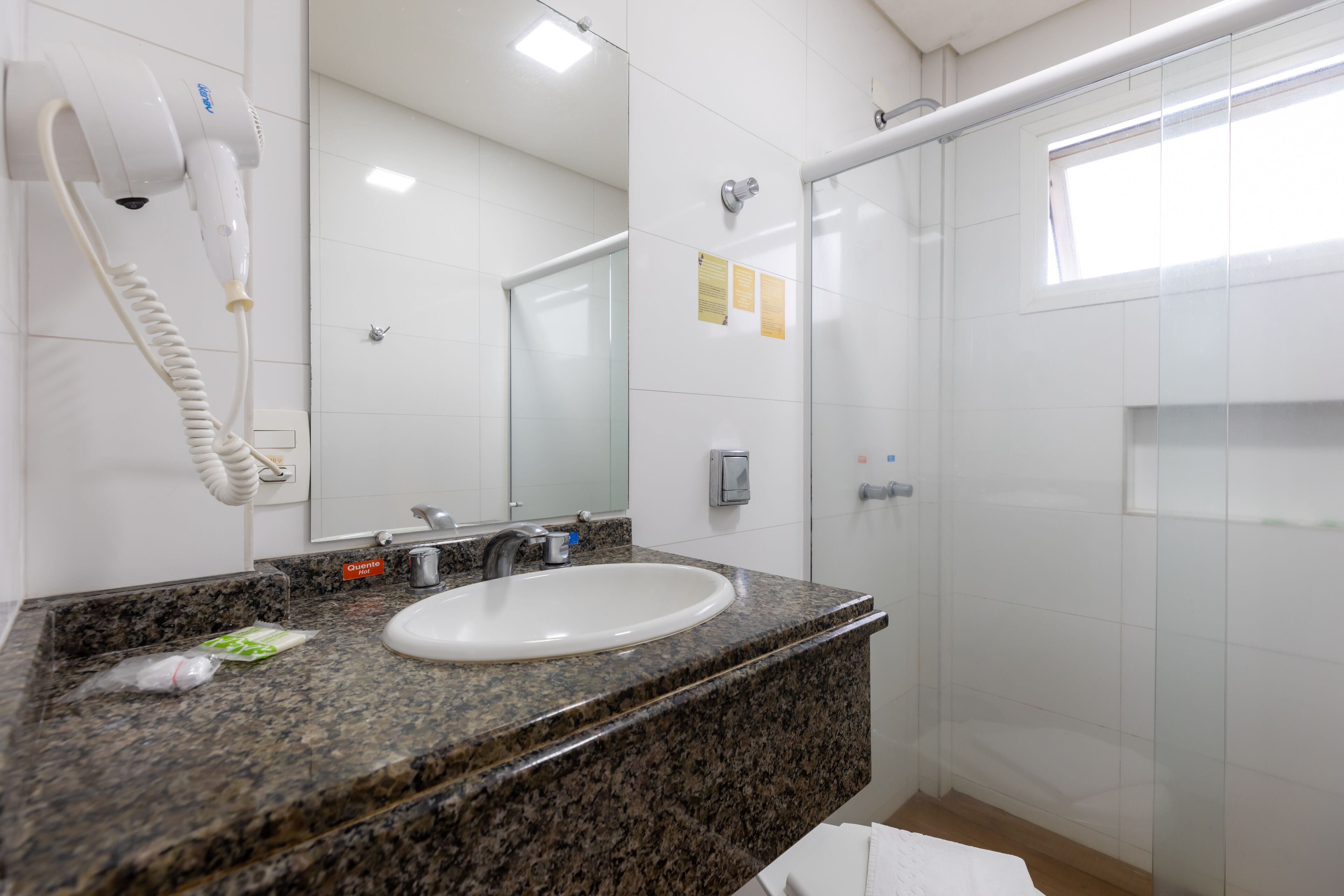 A bathroom with a granite countertop, a white sink, a hairdryer mounted on the wall, a glass shower enclosure, and a small window.