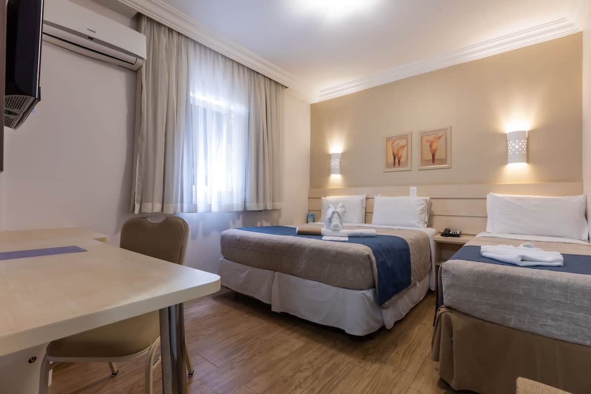 Habitación de hotel con dos camas, un escritorio y una silla. Tiene una ventana con cortinas, un aire acondicionado instalado en la pared y dos apliques.