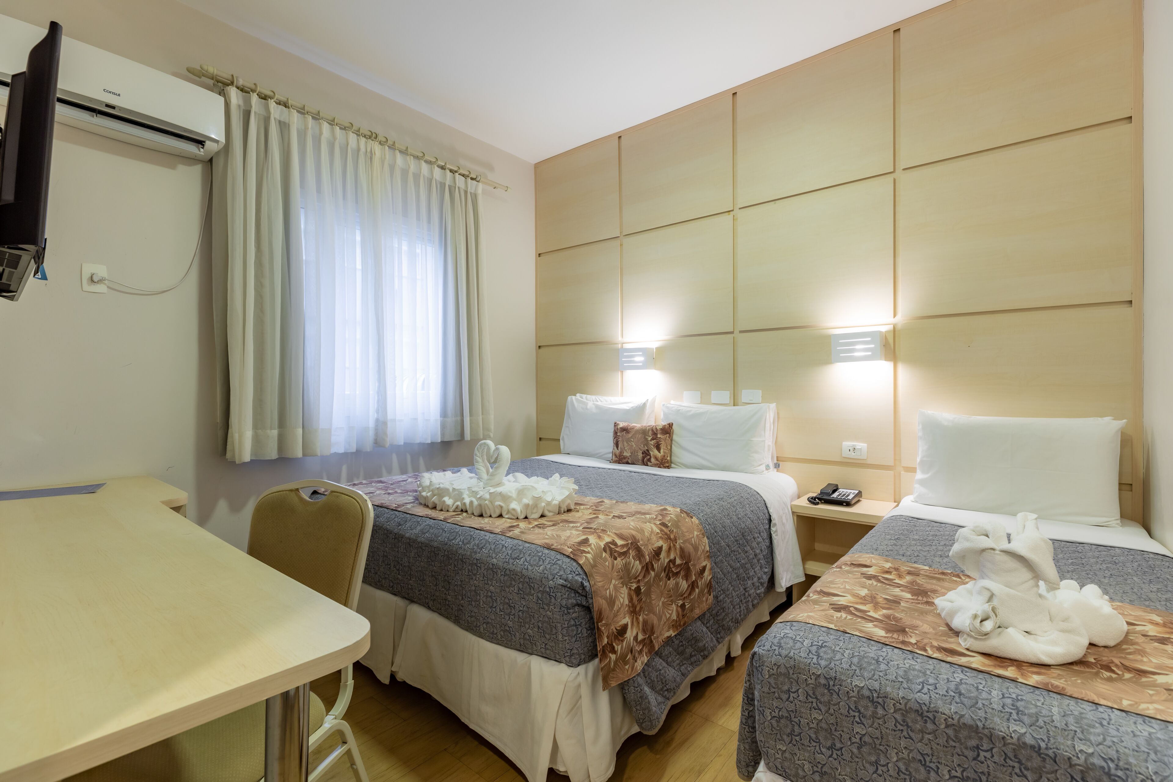 A hotel room with two beds, a desk, and a chair. There is an air conditioning unit on the wall, a curtain, and a bedside table with a phone.
