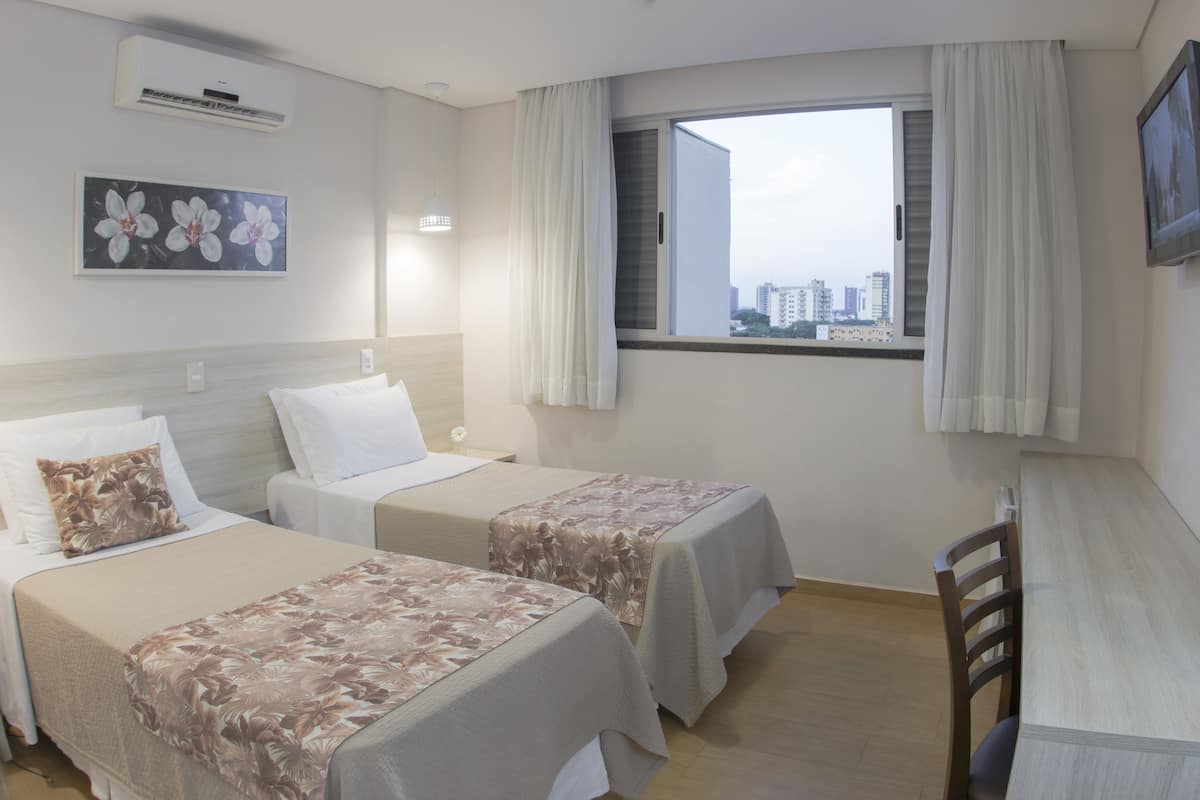 Habitación de hotel con dos camas, un escritorio, un televisor, una ventana con cortinas y vistas a la ciudad.