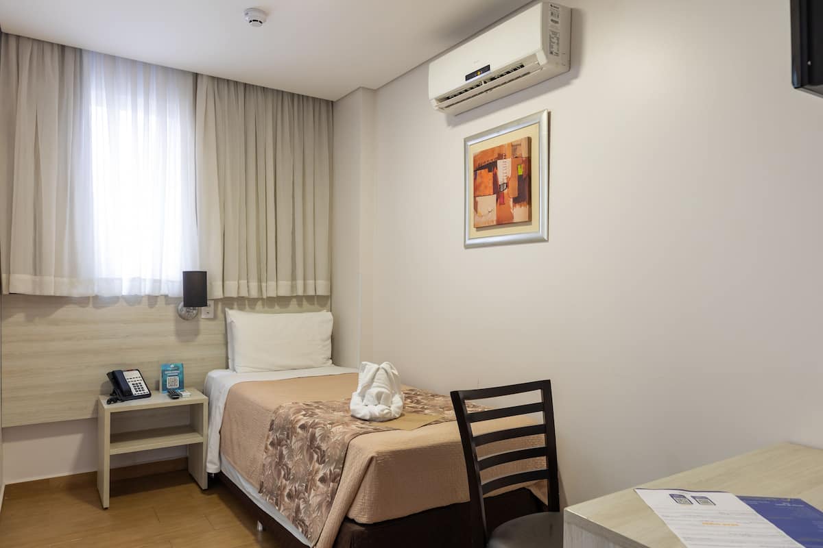 Habitación de hotel con cama, mesita de noche, teléfono, una tarjeta, una silla, aire acondicionado y ventana con cortinas.