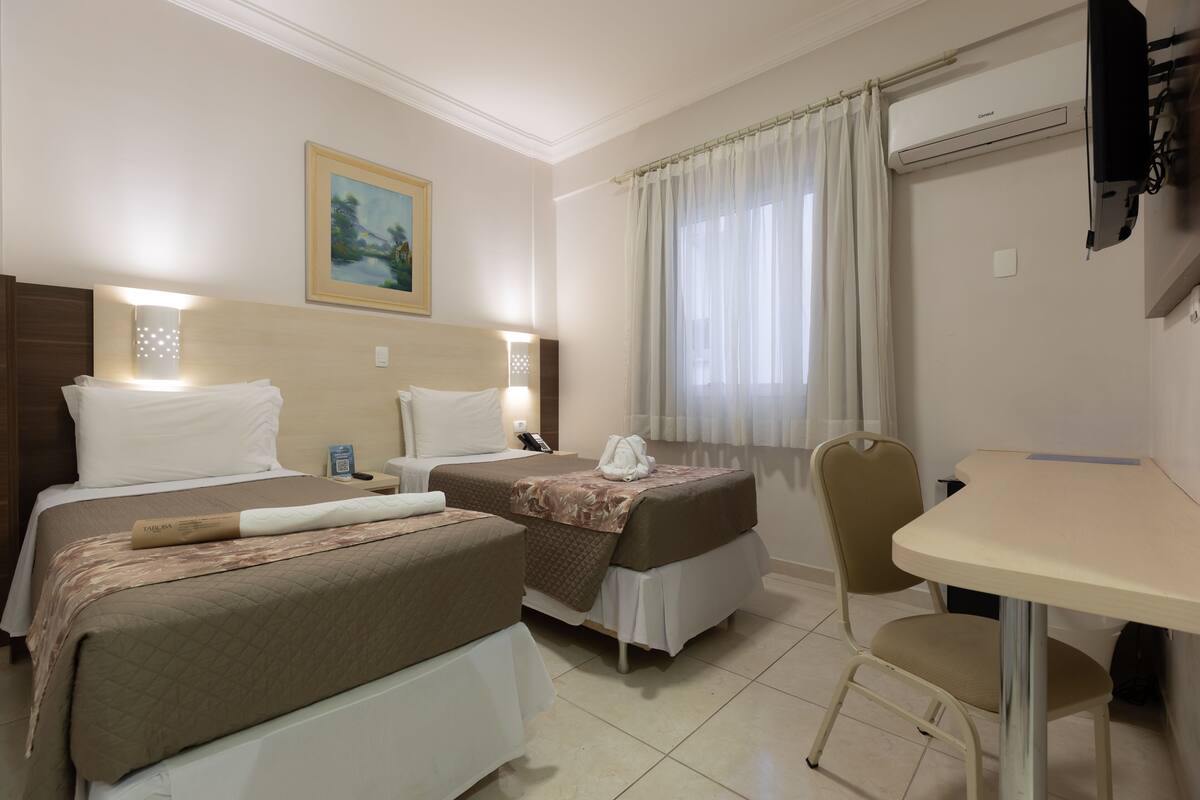 Habitación de hotel con dos camas, un escritorio, una silla, un equipo de aire acondicionado y un cuadro en la pared.