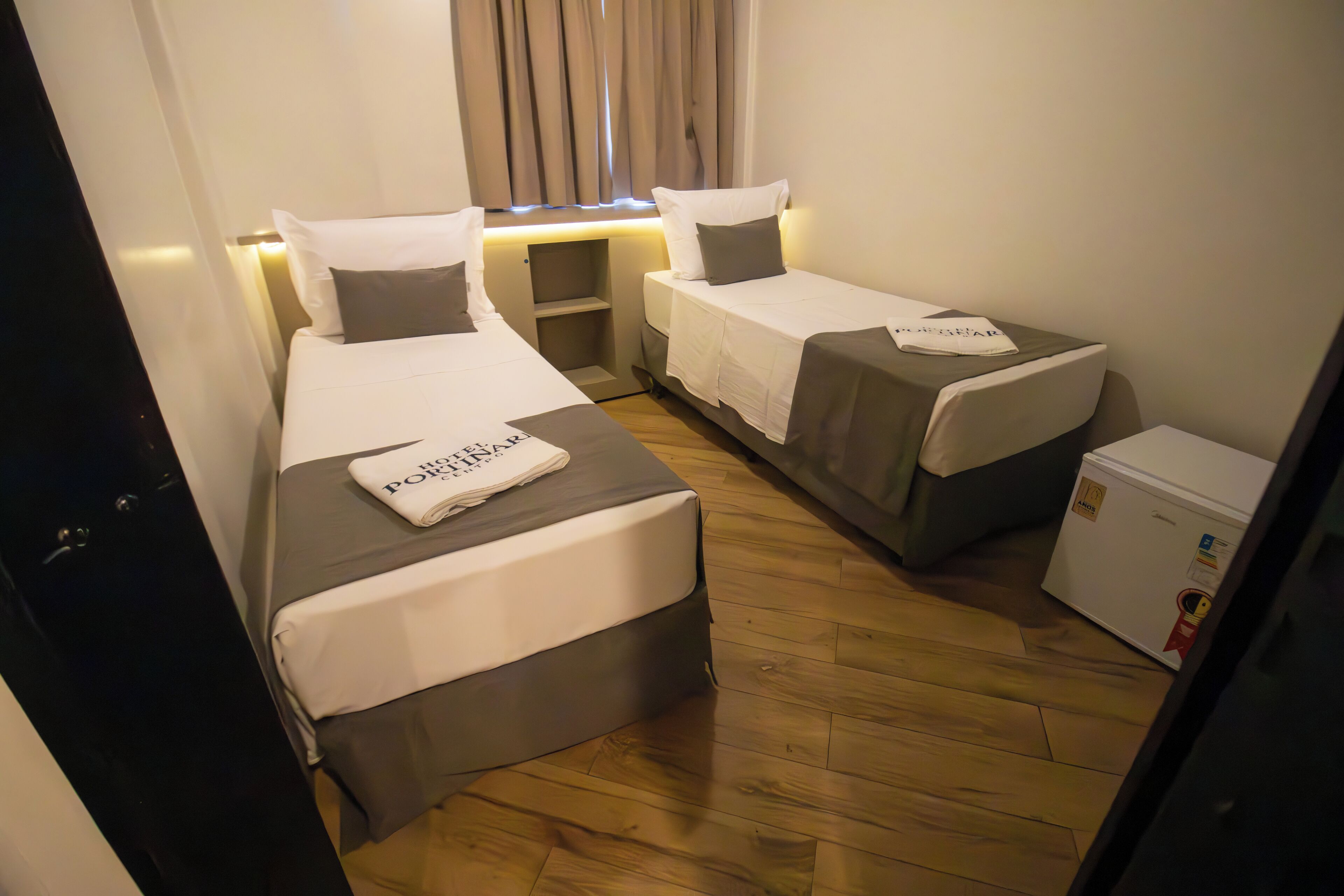 Une chambre d’hôtel avec deux lits simples, une petite table, un réfrigérateur et une fenêtre avec des rideaux.