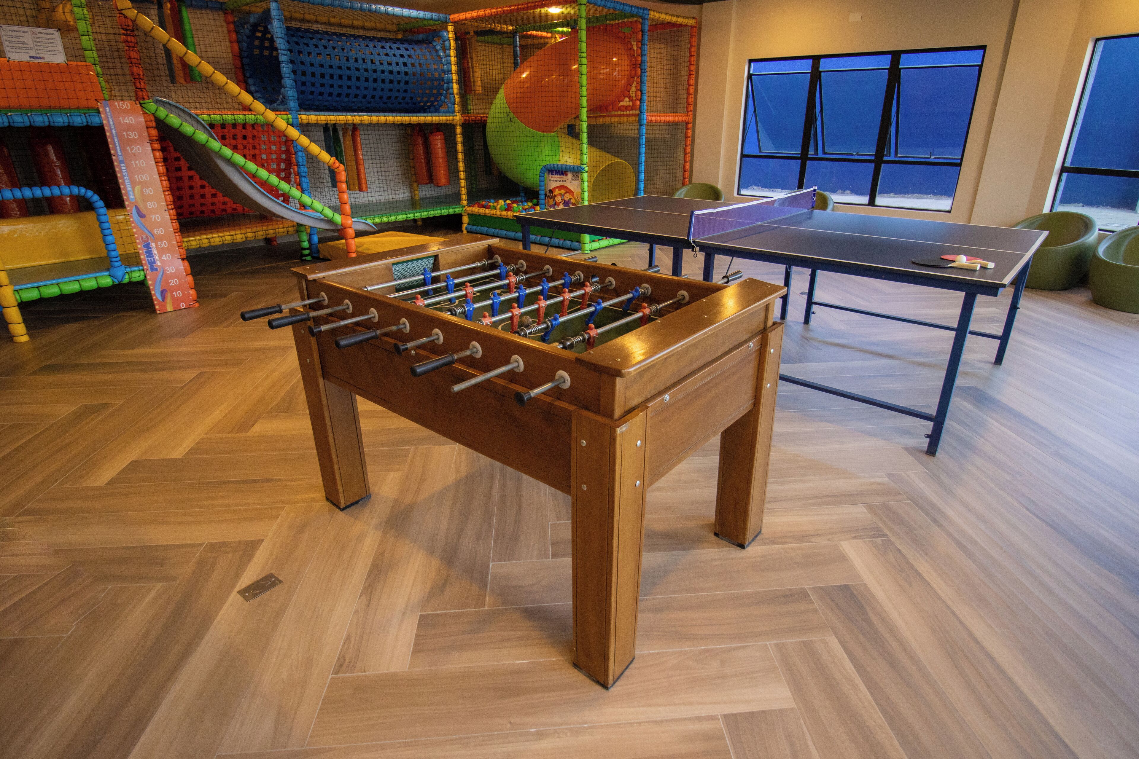 Une table de baby-foot et une table de ping-pong se trouvent dans une pièce dotée d’un parquet en bois disposé en chevrons et de grandes fenêtres.