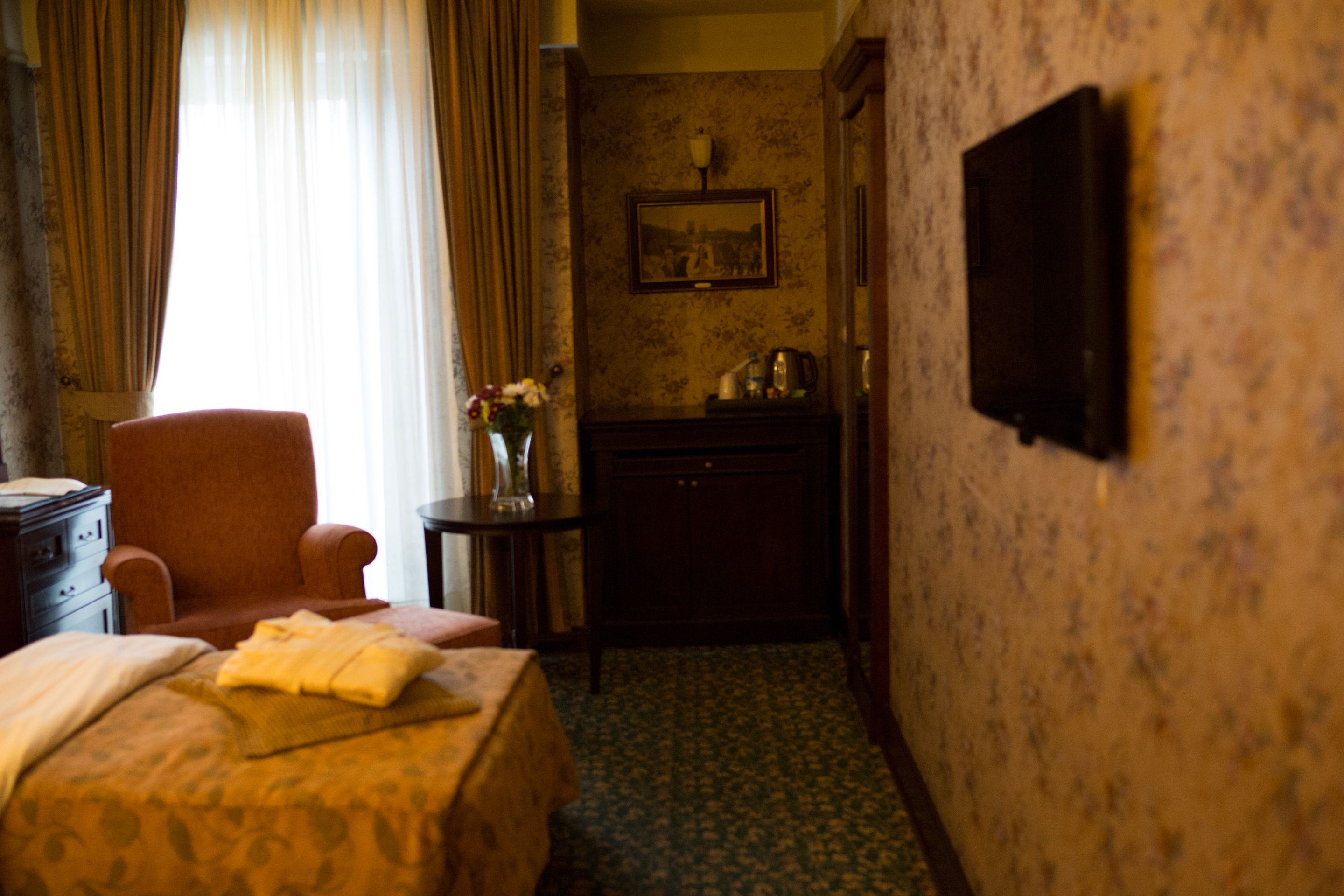 Een hotelkamer met een bed, een stoel, een klein tafeltje met een bloevas, een aan de muur bevestigde televisie en een ingelijste foto.