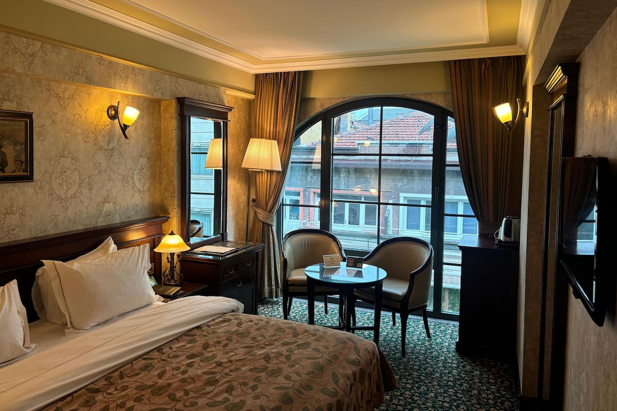 A hotel room with a large bed, a desk with a lamp, a chair, and a small table. There is a window with a view of buildings outside.