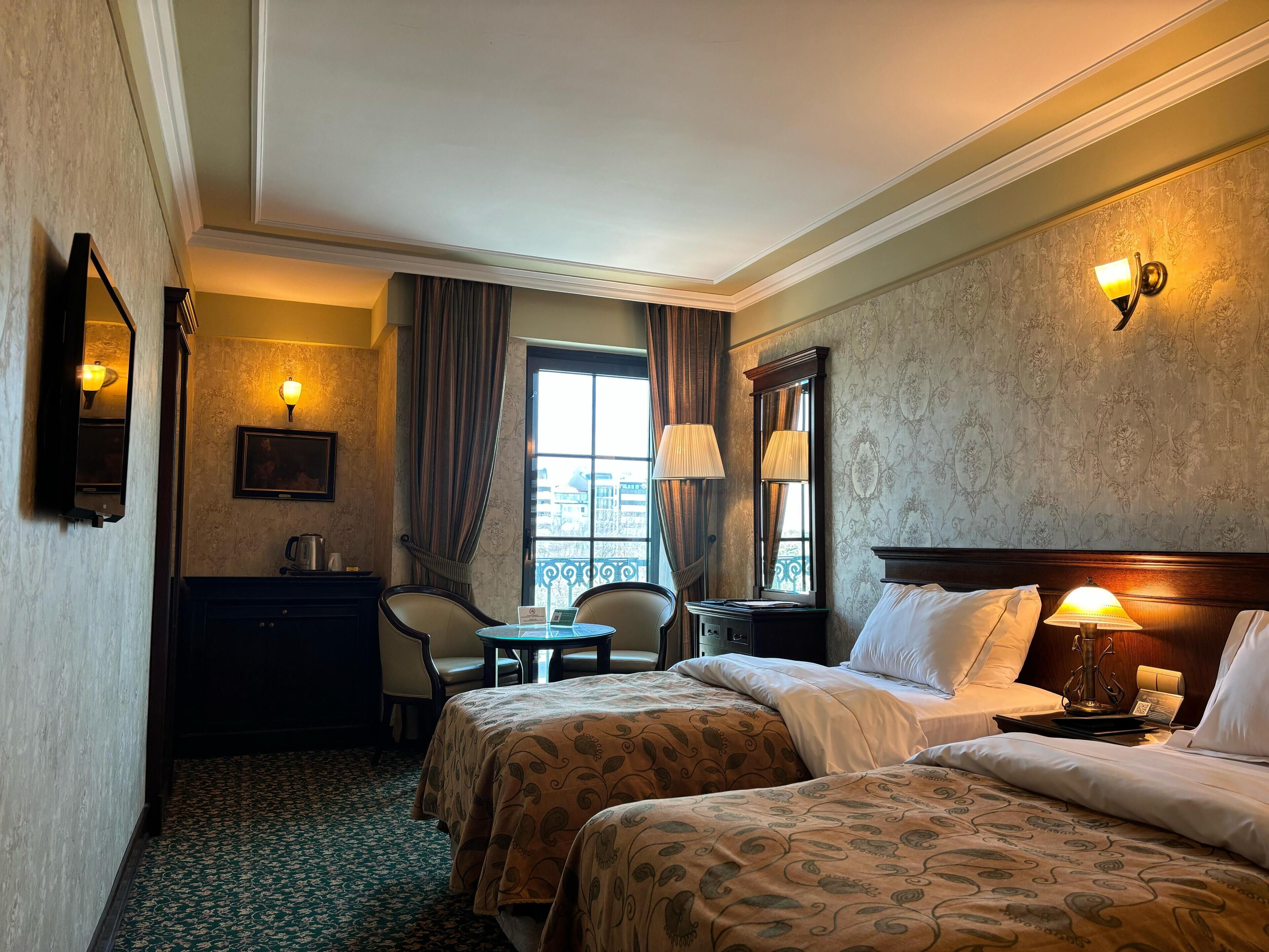 A hotel room with two beds, a desk, a chair, a small table, and a window with curtains.