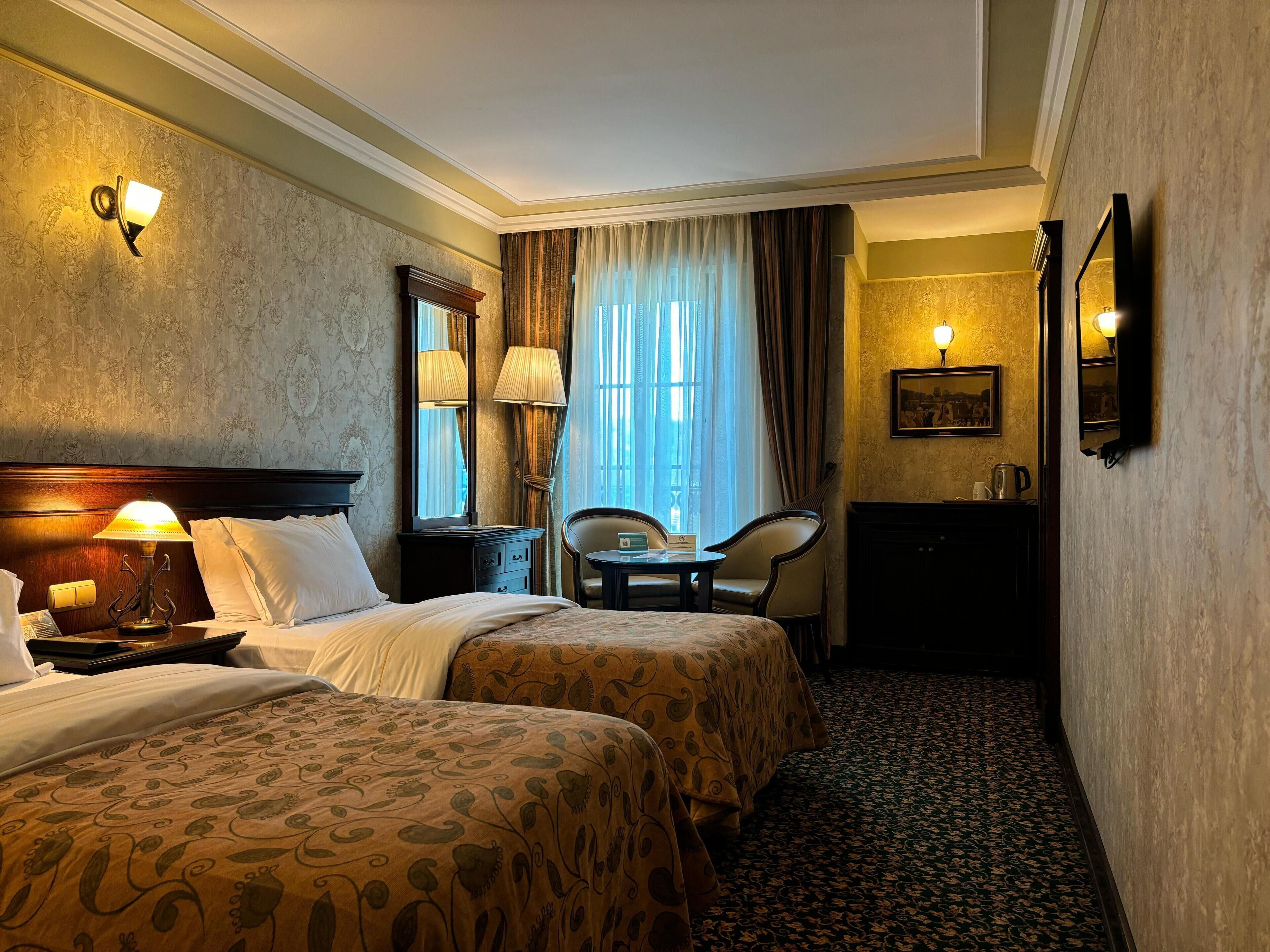 Une chambre d’hôtel avec deux lits, un bureau, une chaise, une table de chevet, une lampe, un miroir et un tableau au mur.