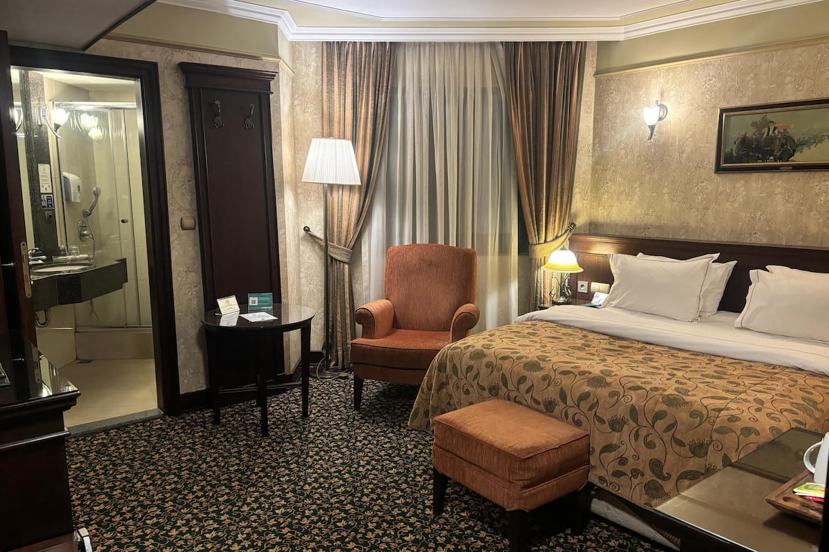 A hotel room with a bed, a chair, a small table, a desk, and a bathroom visible through an open door.