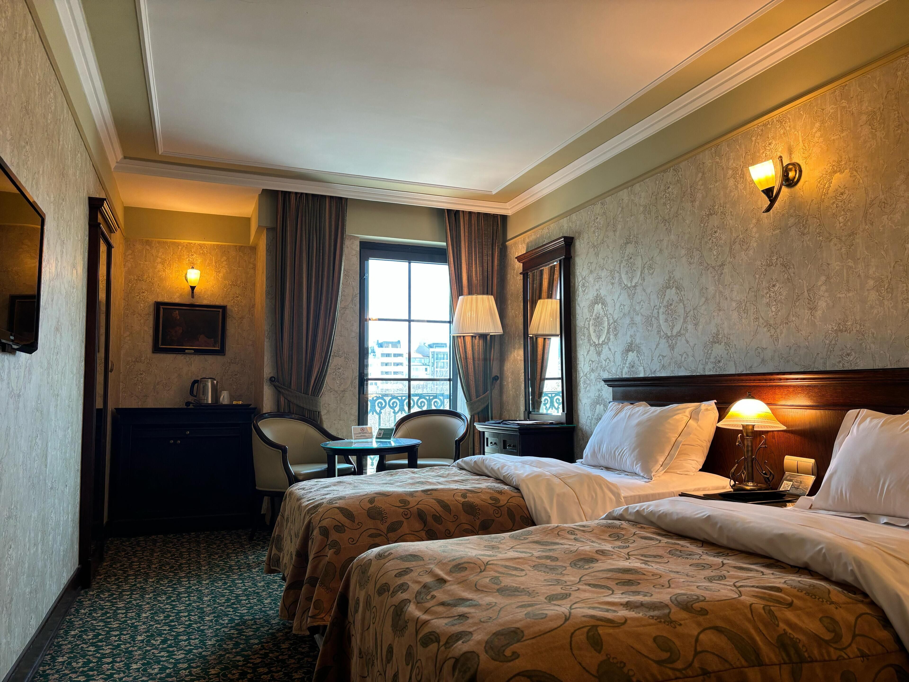 A hotel room with two beds, a sitting area with a chair and table, a large window with curtains, and a wall-mounted lamp.