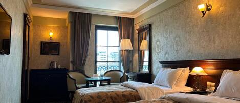 A hotel room with two beds, a sitting area with a chair and table, a large window with curtains, and a wall-mounted lamp.