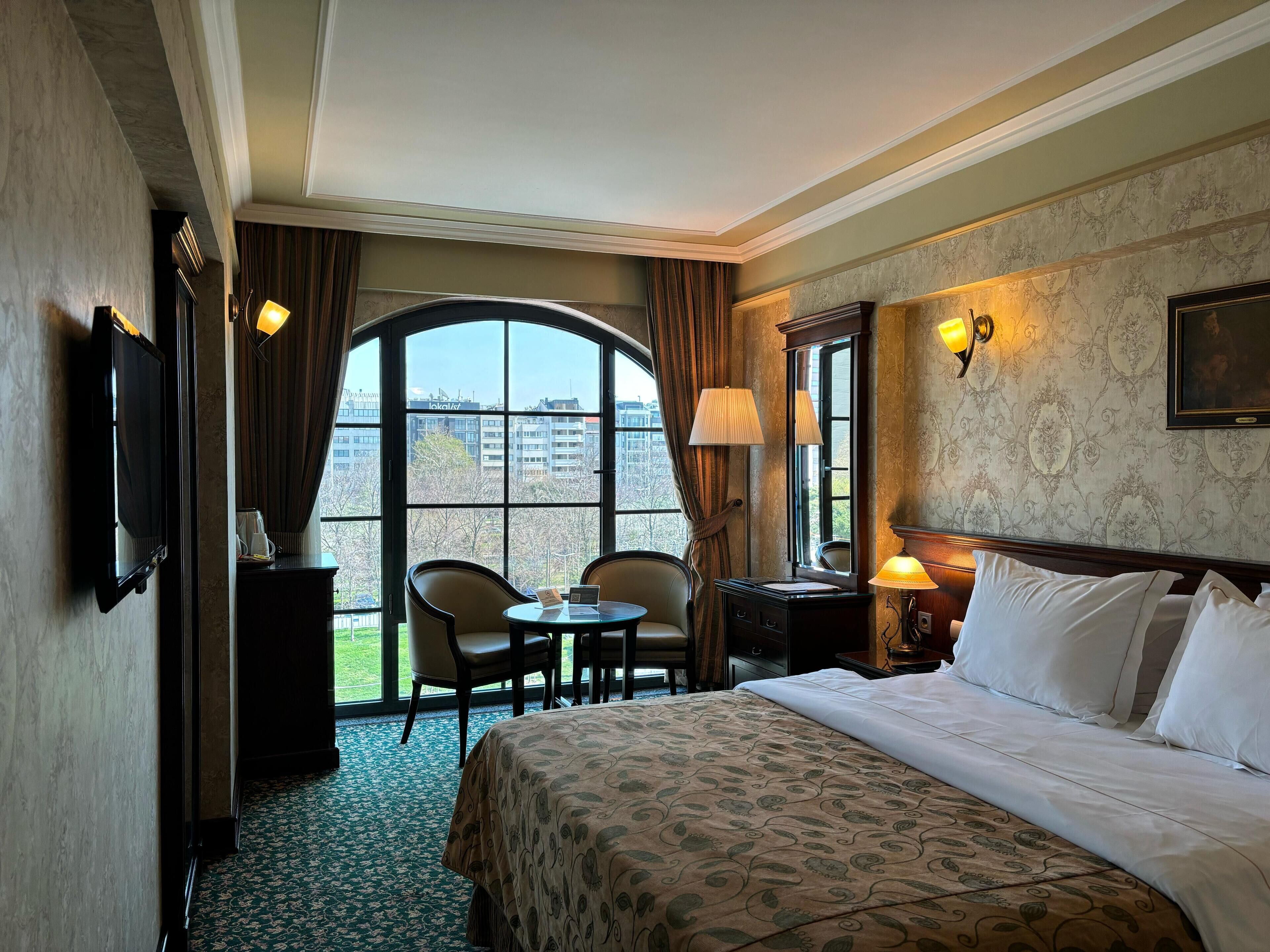 A hotel room with a large bed, two chairs, a small table, a TV, and a window with a view of buildings.