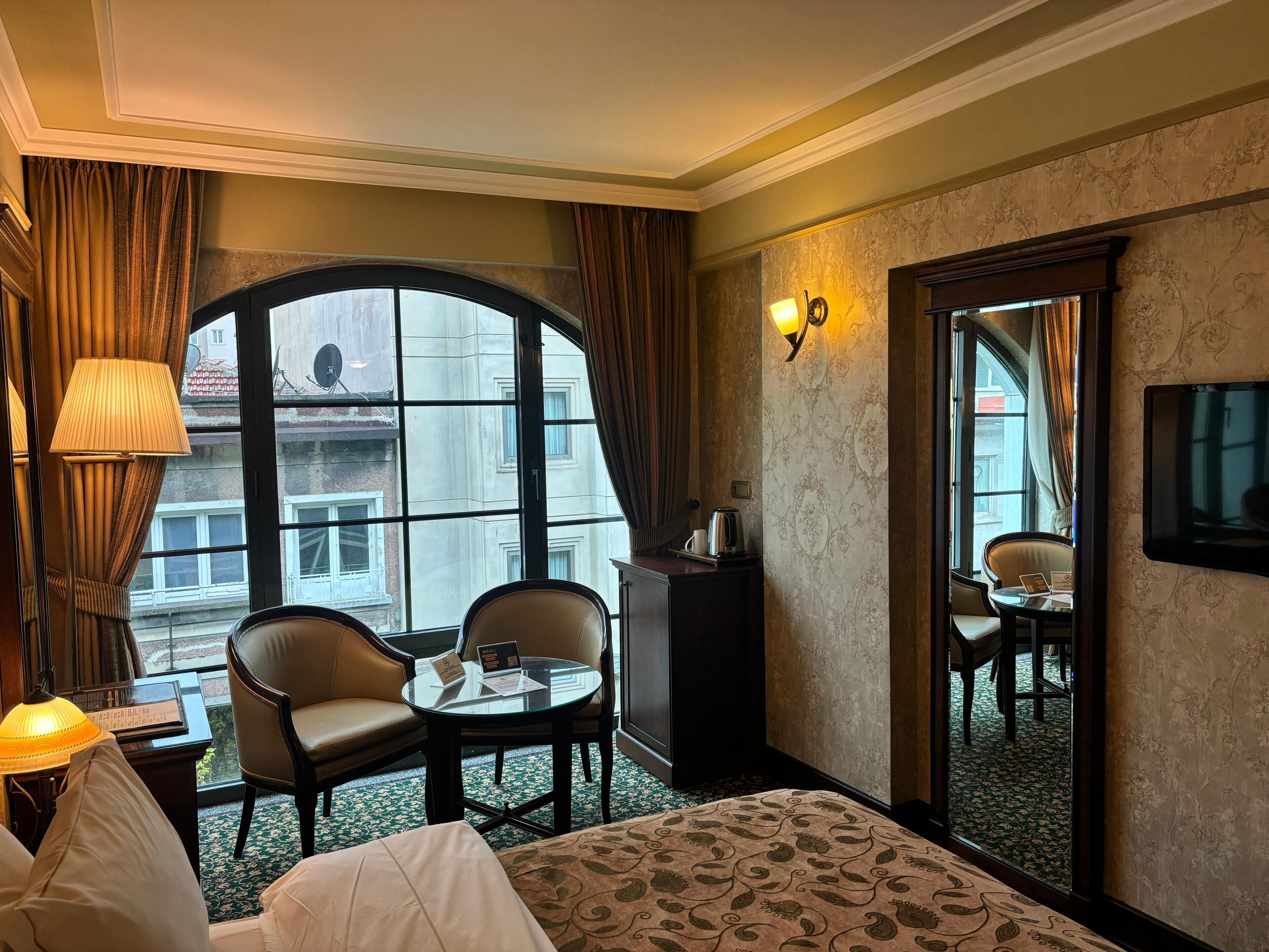 A hotel room with a large arched window, two chairs, a small table, a bed, a bedside table with a lamp, a mirror, and a TV on the wall.