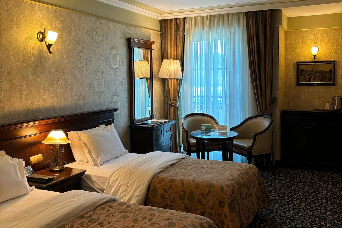 A hotel room with two beds, a desk with a chair, a lamp, a nightstand, and a window with curtains.