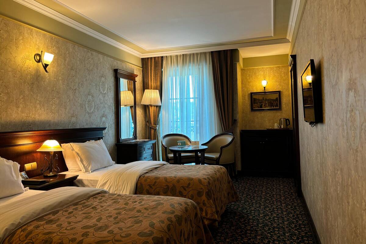 A hotel room with two beds, a desk, a chair, a lamp, and a window with curtains.