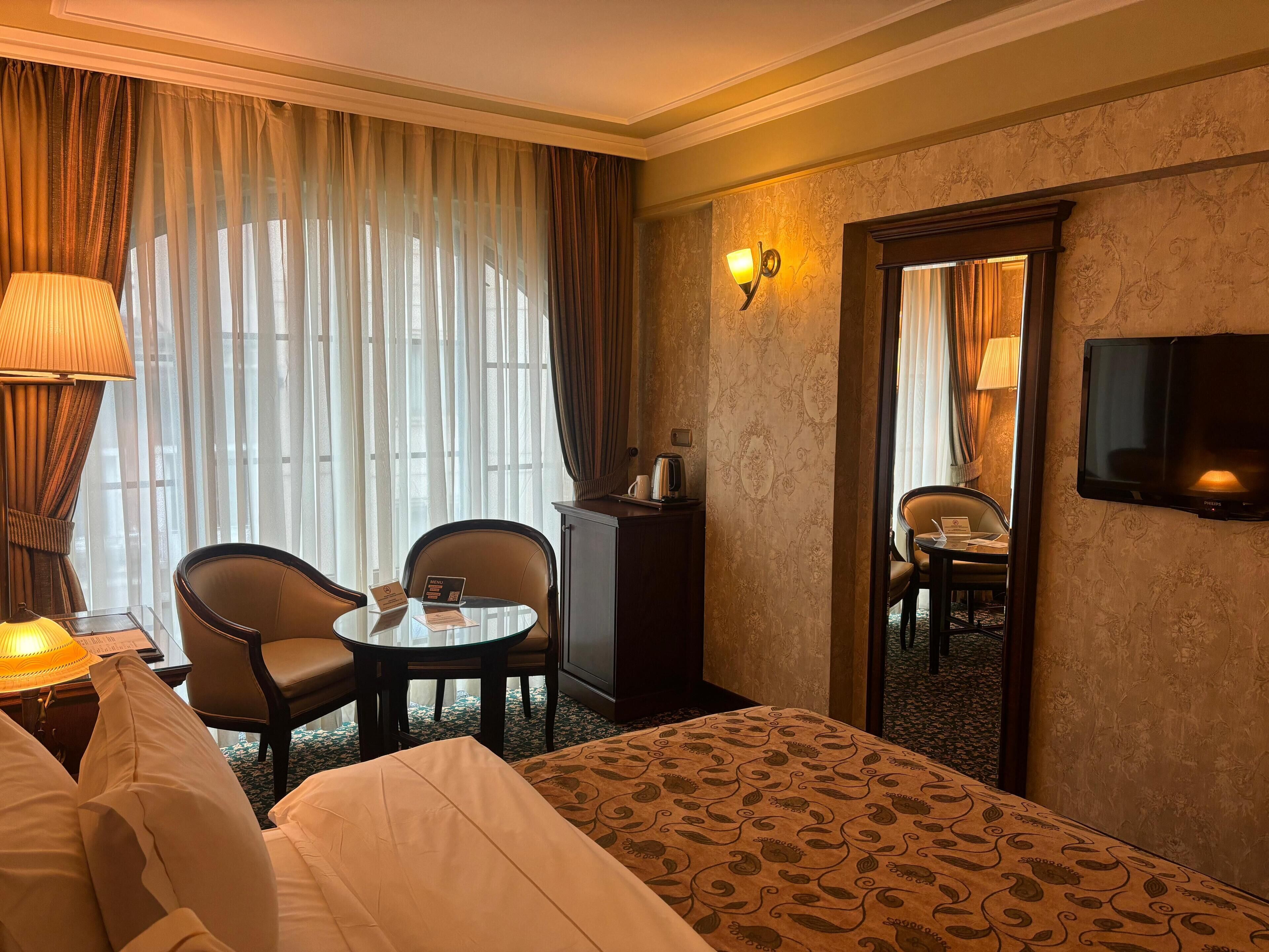 A hotel room with a bed, a desk with a lamp, two chairs, a small table, a television, and a large window with curtains.