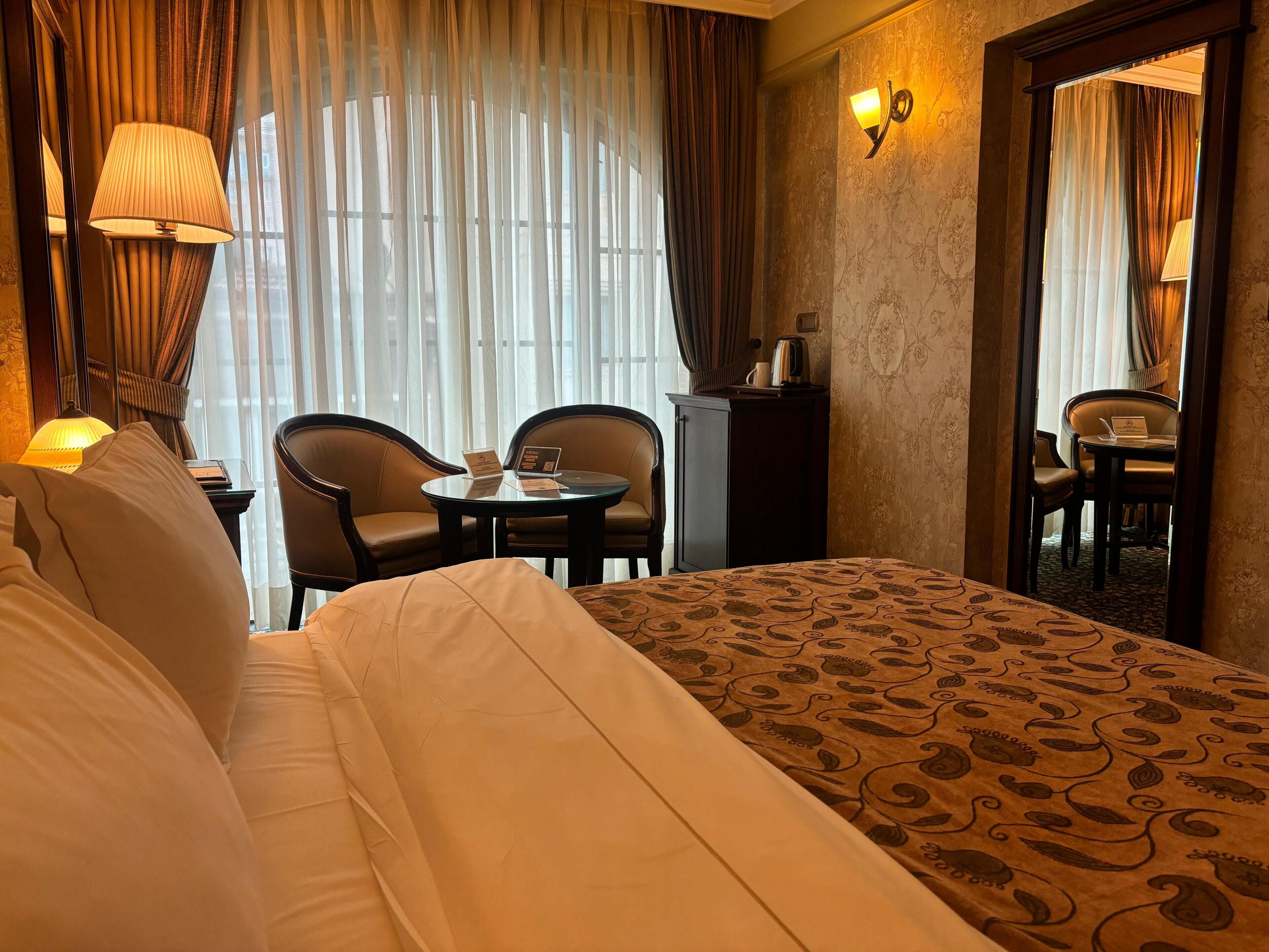 A hotel room with a large bed, two armchairs, a small round table, a minibar, and a large mirror.