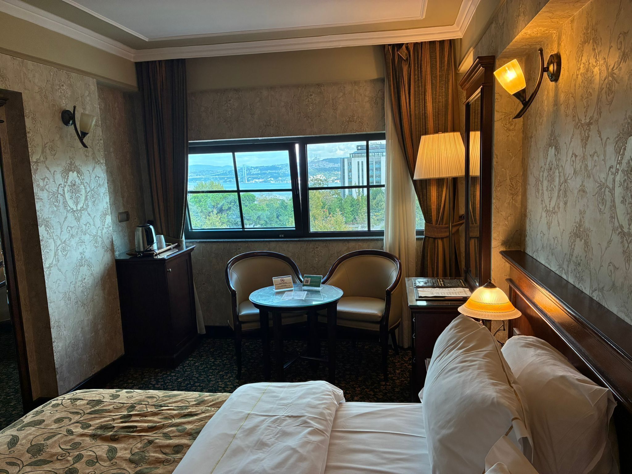 A hotel room with a bed, a desk, a chair, a table, a lamp, and a window with a view.