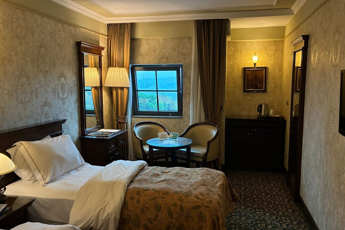 A hotel room with a bed, a desk with a lamp, a chair, a small table, a window with curtains, and a painting on the wall.