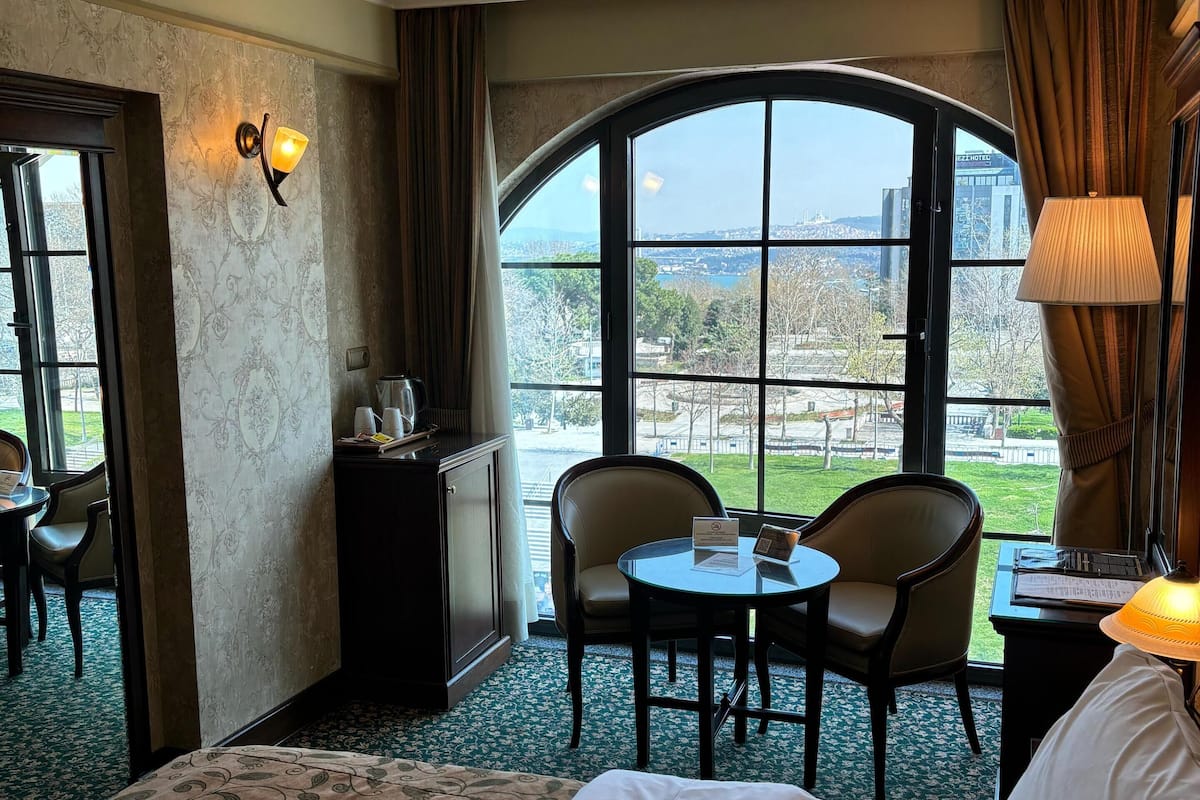 A hotel room with a large bed, a desk with a chair, a small table, and a view of the city through a large window.