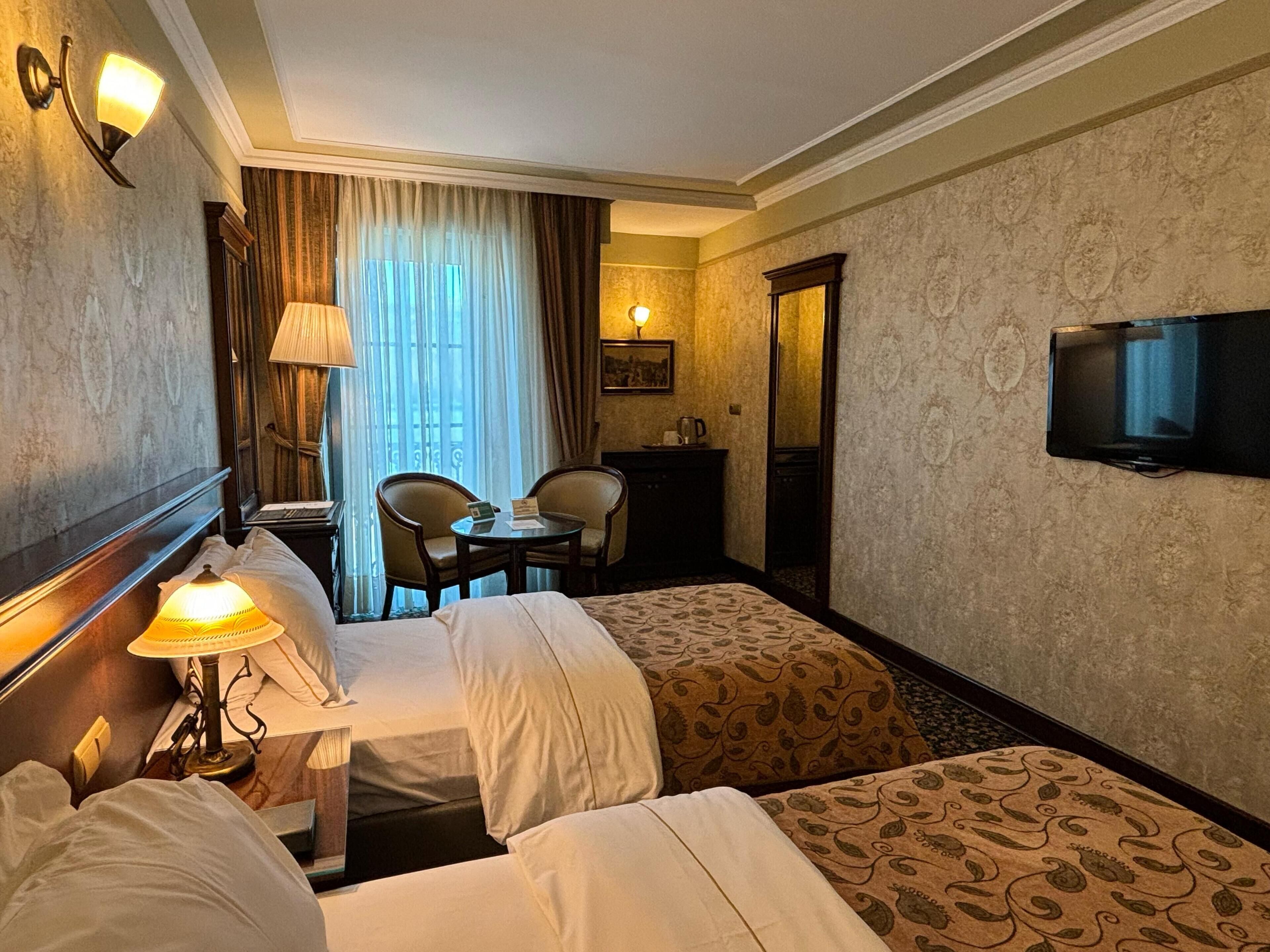 A hotel room with a bed, two armchairs, a small table, a desk, a lamp, a television, and a window with curtains.