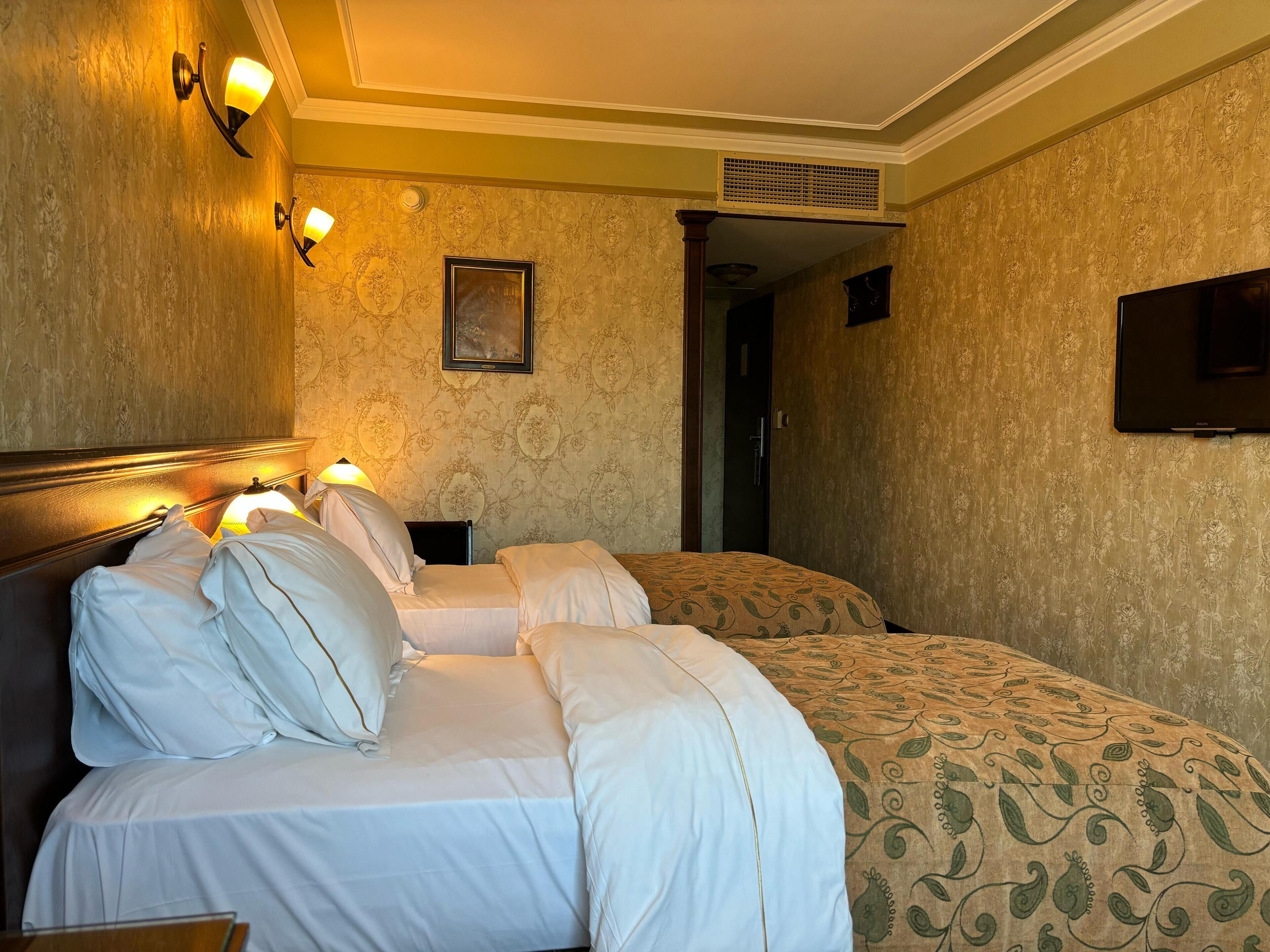 A hotel room with two beds, a headboard, wall sconces, a framed picture, and a television.