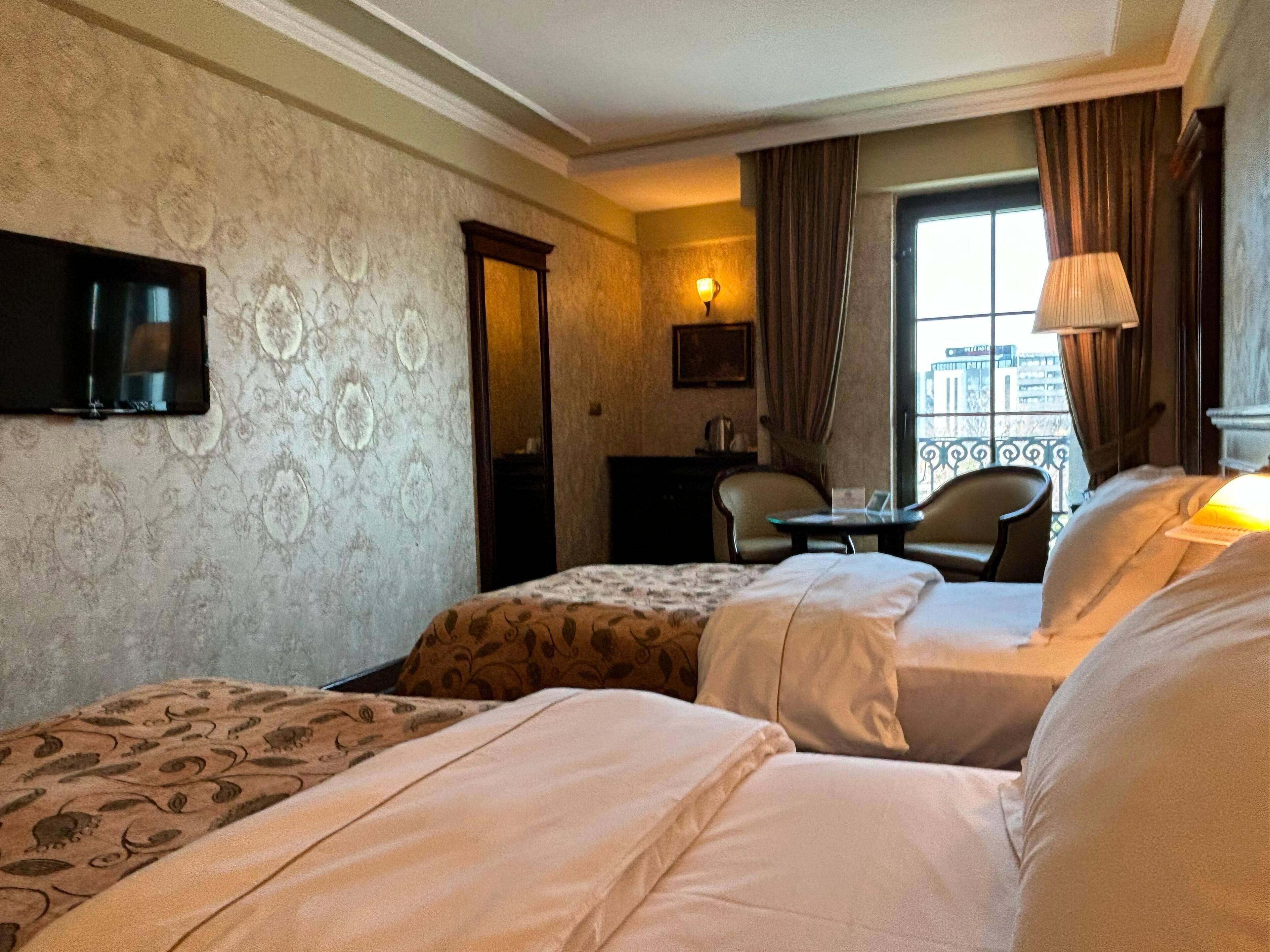A hotel room with two beds, a small table, a chair, a TV mounted on the wall, and a window with curtains.