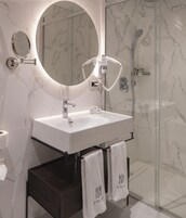Un bagno moderno con un lavandino bianco, uno specchio rotondo e un soffione doccia manuale. Ci sono due asciugamani su un portascopevoli con il nome dell'hotel.