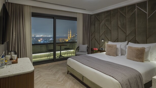 Ein Hotelzimmer mit einem großen Bett, einem Schreibtisch und Blick auf eine beleuchtete Stadt bei Nacht.
