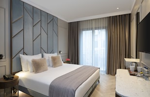 Habitación de hotel con una cama grande, una mesita de noche con lámpara, un televisor de pantalla plana y vistas a la ciudad a través de amplios ventanales.