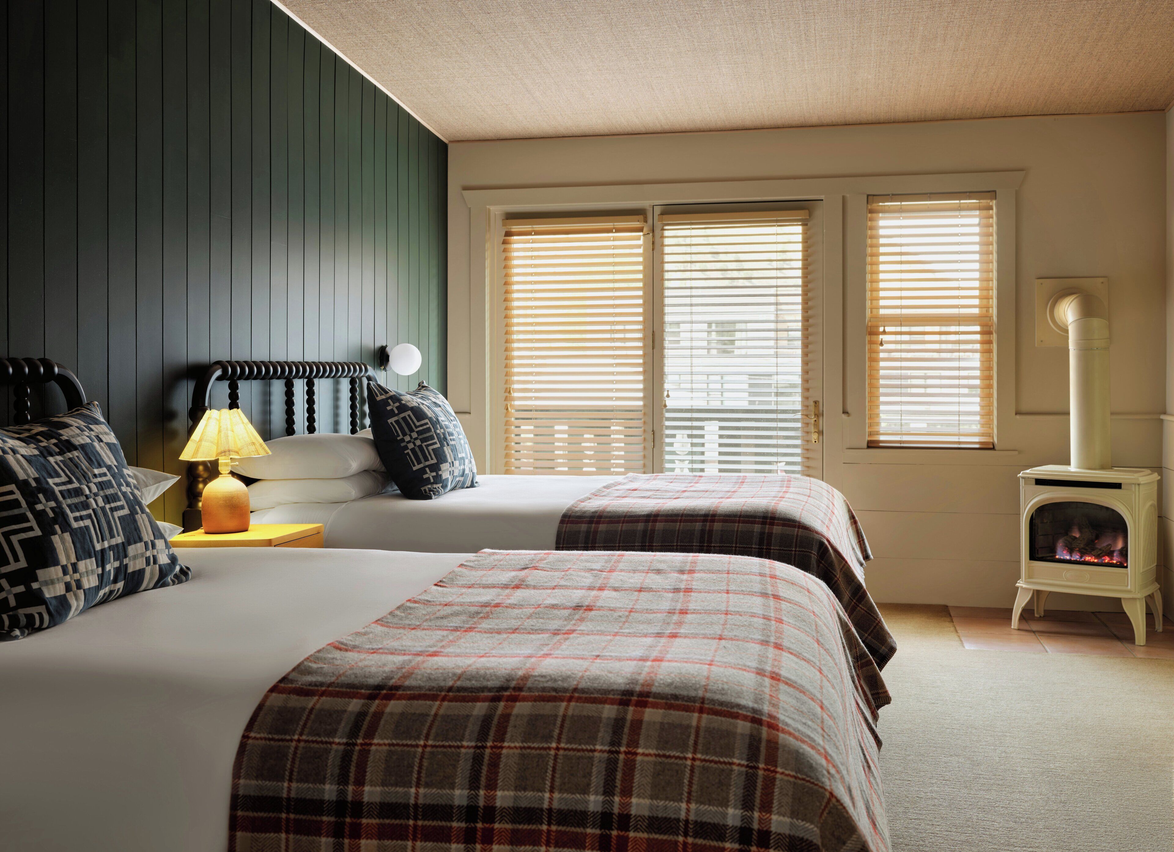 Une chambre avec deux lits, une table de chevet, une lampe, une fenêtre avec des stores et un radiateur blanc.