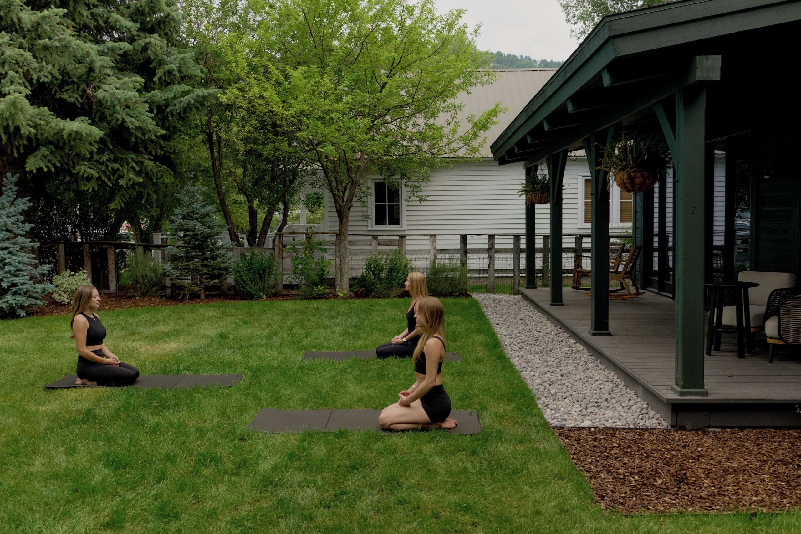 Two individuals seated on yoga mats in a grassy outdoor area with a building in the background.