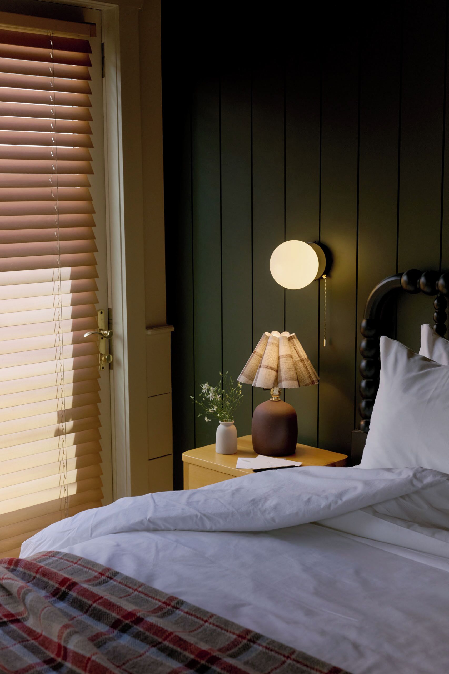 Un dormitorio con cama, una mesita de noche con lámpara, un jarrón con flores y una ventana con persianas.